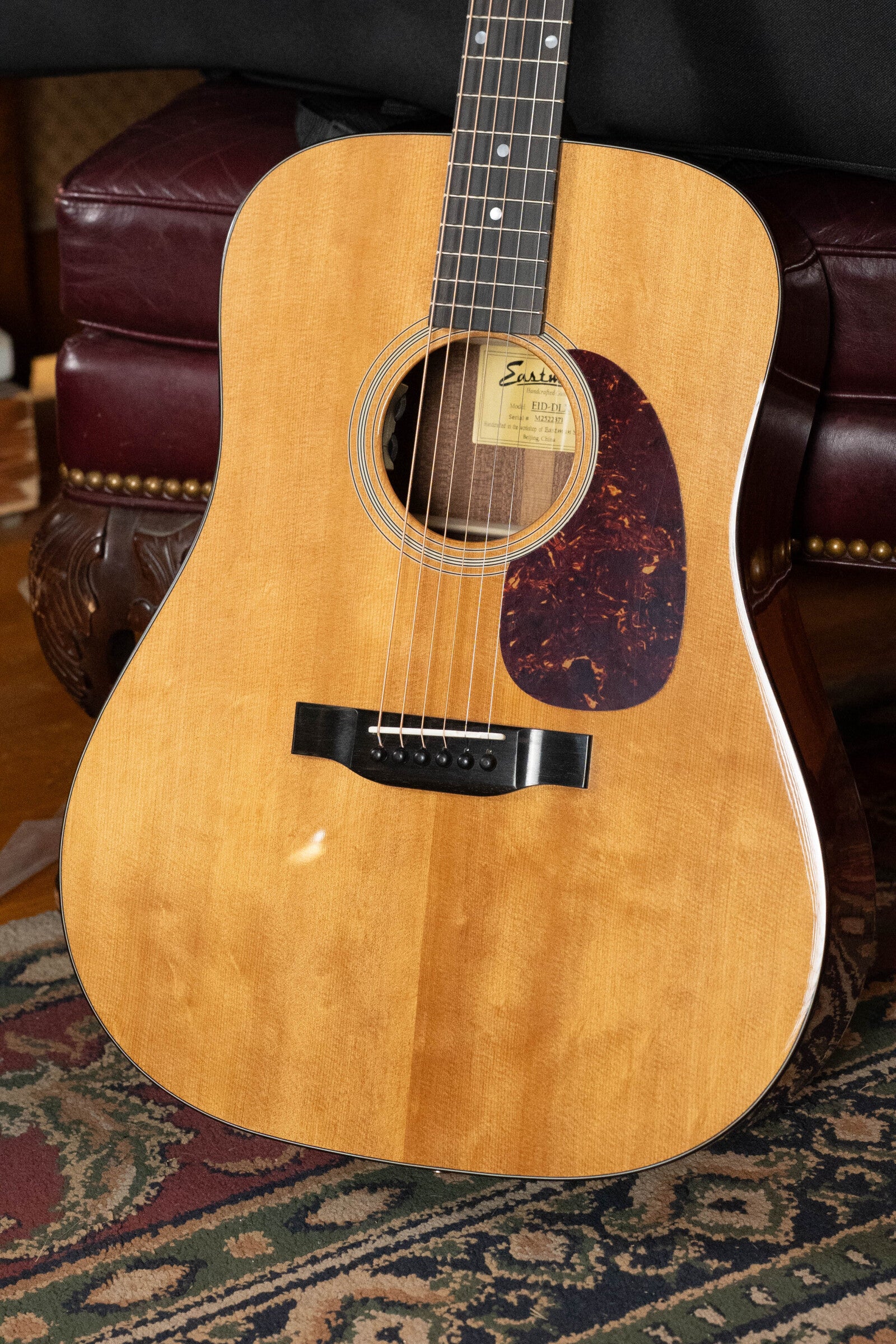 An Eastman Guitars E1D-DLX Sitka/Sapele Dreadnought w/Fishman Pickup (#2371), featuring a natural finish and tortoiseshell pickguard, sits on a patterned rug before a maroon leather chair.