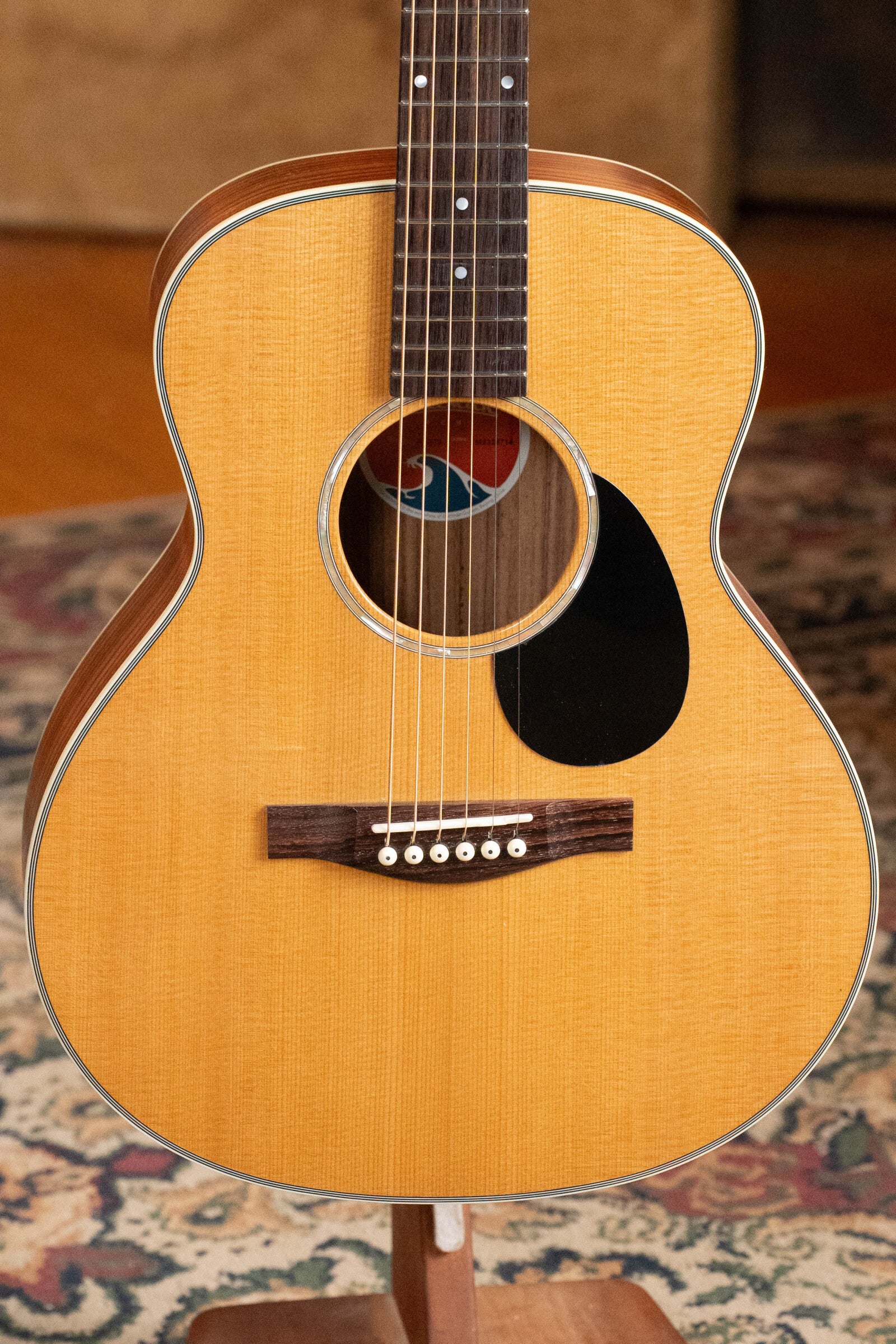 Close-up of the Eastman Guitars PCH2-TG Travel Acoustic Guitar (#9714) with thermo-cured solid Sitka top, laminated rosewood body, colorful sound hole label, six strings, dark pickguard, and part of a patterned rug in the background.