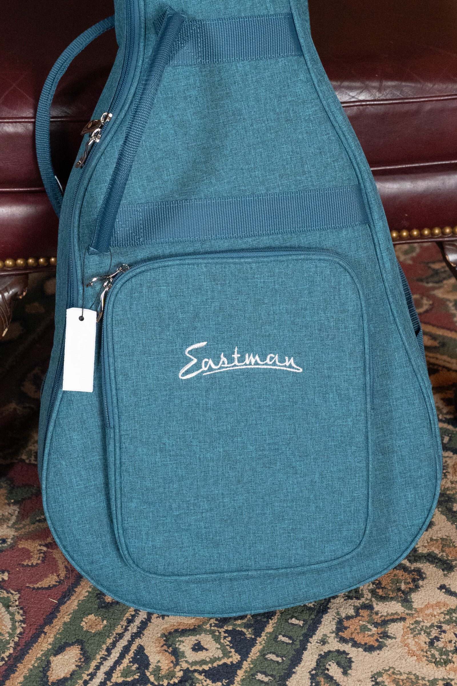 An Eastman Guitars PCH2-TG Thermo-Cured Solid Sitka/Laminated Rosewood Travel Acoustic Guitar #9714 case with a zipper pocket and white logo sits on a patterned rug near a dark red chair.