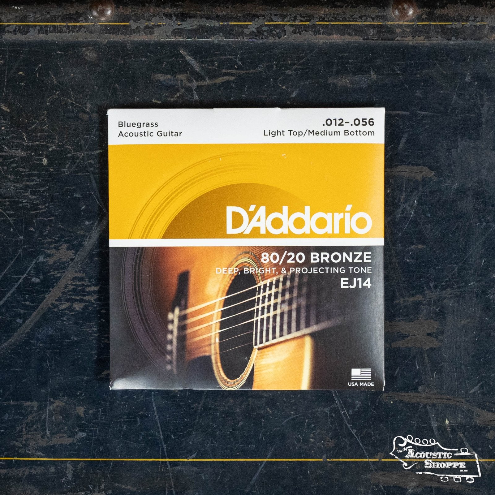 A pack of D'Addario EJ14 Light Top/Medium Bottom Bluegrass, 80/20 Bronze Acoustic Guitar Strings (12-56) sits on a dark surface. The packaging shows an acoustic guitar—perfect for bright, bluegrass-inspired tone seekers.