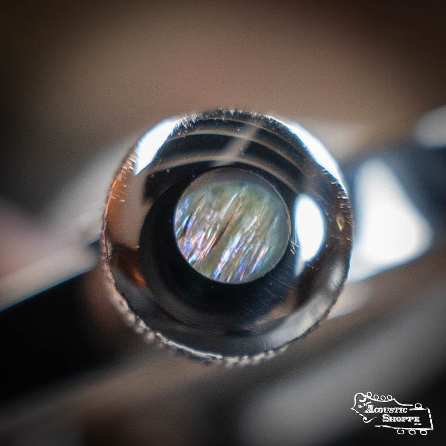 Close-up of the Elliott G34E Push Button Elite Capo for guitar (1 3/4 w/ 16 radius), featuring a shiny abalone inlay screw. Blurred reflections and the Acoustic Shoppe logo appear in the bottom right corner.