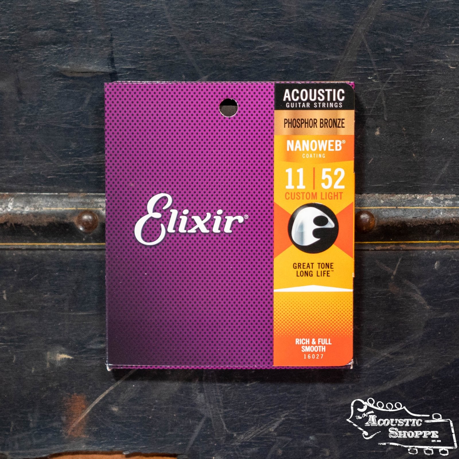 A box of Elixir 16027 Acoustic Nanoweb Custom Light 11-52 guitar strings by Elixir sits on a dark textured surface, featuring purple and orange packaging with detailed product info—ideal for acoustic guitars.
