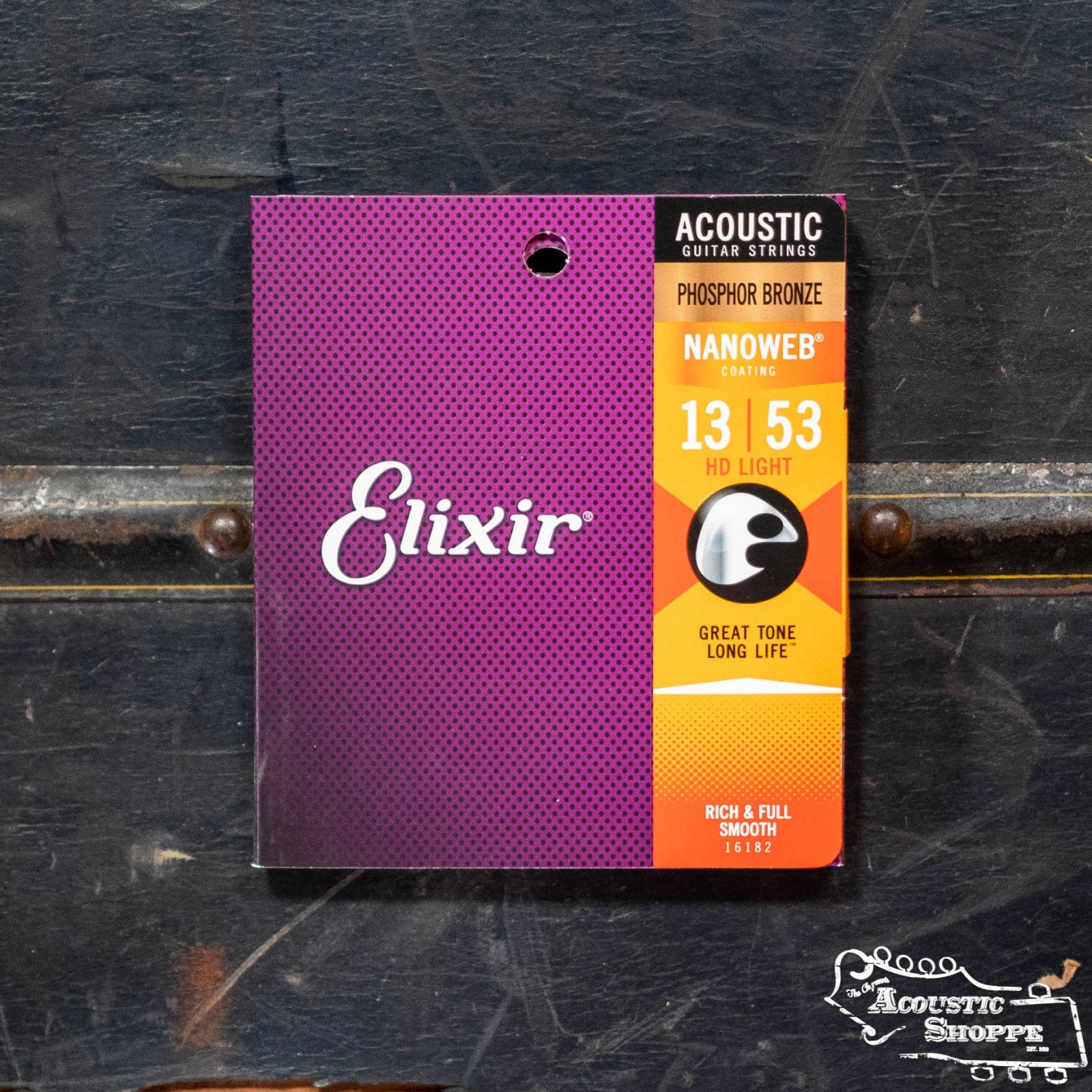 The Elixir 16182 Acoustic HD Light Phosphor Bronze 13-53 guitar strings pack is shown on a dark wooden surface. The packaging features Elixir branding in purple and orange, ideal for acoustic music stores.