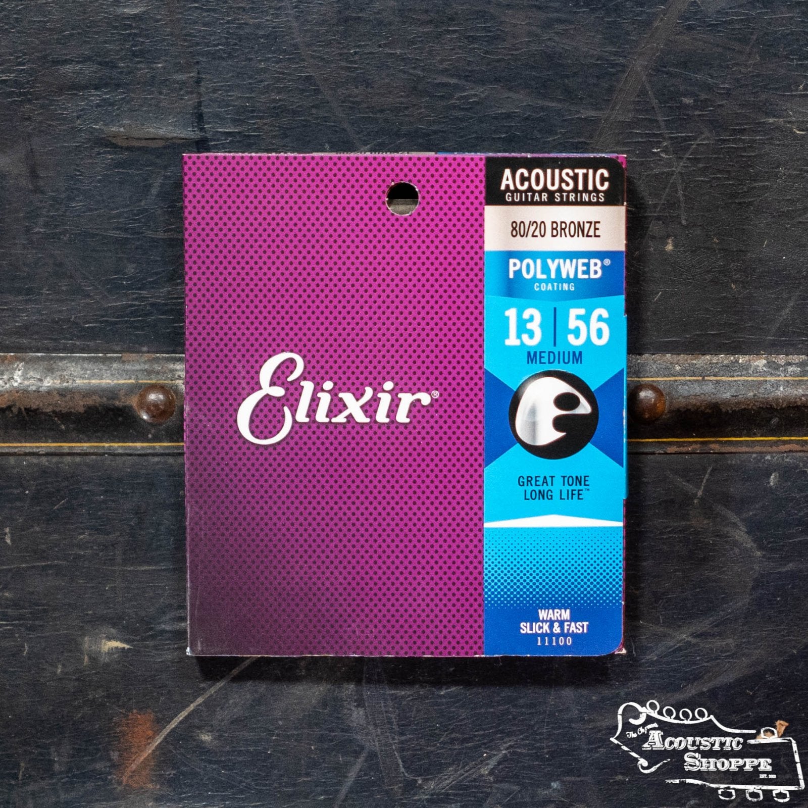 A pack of Elixir 11100 Acoustic 80/20 Bronze Strings with POLYWEB Coating (13-56) by Elixir is shown in purple packaging, displaying branding and product details on a dark wooden surface.
