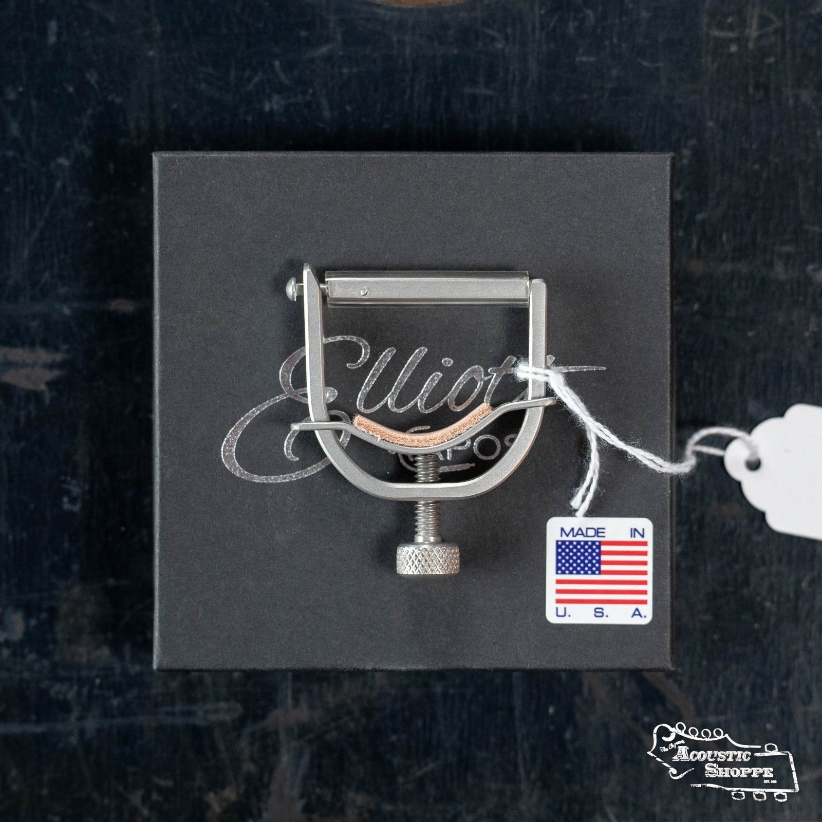 The Elliott B450I Integrity Banjo Capo (Top Bar 1.400-1.450" Flat Radius) by Elliott Capos is shown in silver atop a black Elliott box, with a Made in U.S.A. tag—ideal for music stores or acoustic instrument fans on a dark textured background.