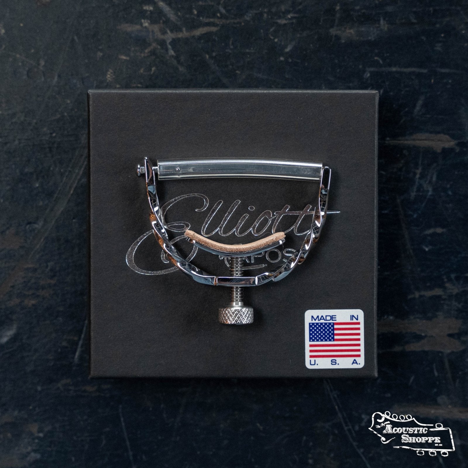 An Elliott Capos G34E-TW Elite Guitar Capo (Twist Stainless Steel, 12" Radius) is displayed on a black box marked "Elliott," with an American flag sticker stating Made in U.S.A., all set atop a dark, textured surface.
