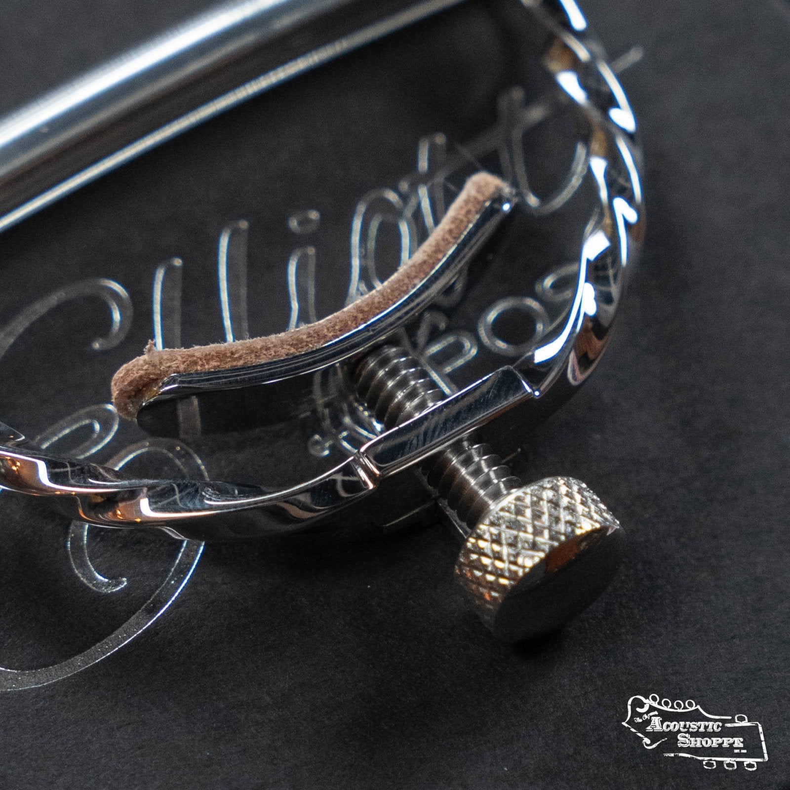 Close-up of the Elliott Capos G34E-TW Elite Guitar Capo (Twist, Stainless Steel, 16 Radius) with brown felt strip, on a black Elliott Capos-engraved surface. The Acoustic Shoppe logo appears in the bottom right corner.