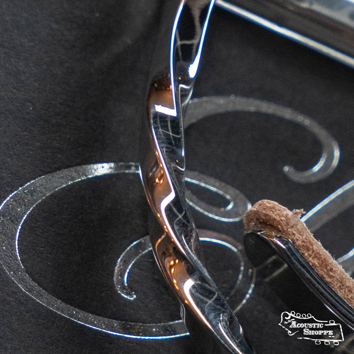 Close-up of the Elliott Capos G34E-TW Elite Guitar Capo, a shiny stainless steel capo with a textured brown tip, resting on a black surface with white decorative patterns and an Acoustic Shoppe logo.
