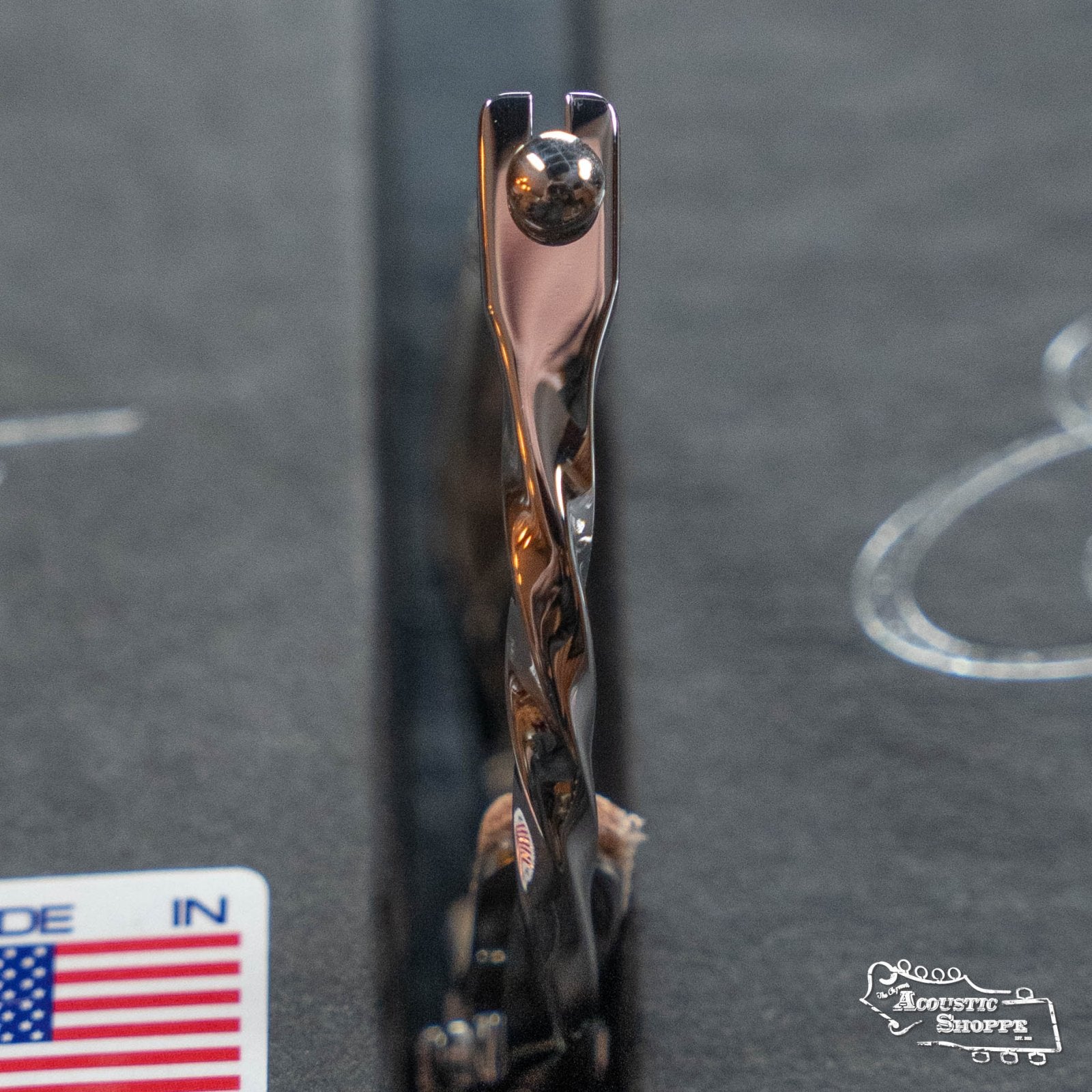 Close-up of an Elliott G34E-TW Elite Guitar Capo—Twist Stainless Steel 12" Radius by Elliott Capos—resting in a hard case, with a partial Made in USA sticker and Acoustic Shoppe logo visible.