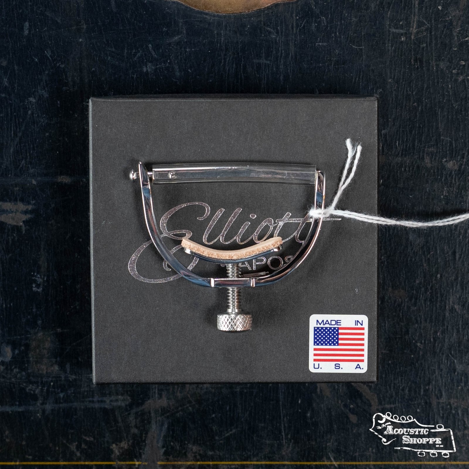 A polished Elliott G78E Push Button Elite Capo by Elliott Capos sits on a black box with a Made in USA sticker and tied string—an ideal accessory for boutique guitar and acoustic instrument enthusiasts.