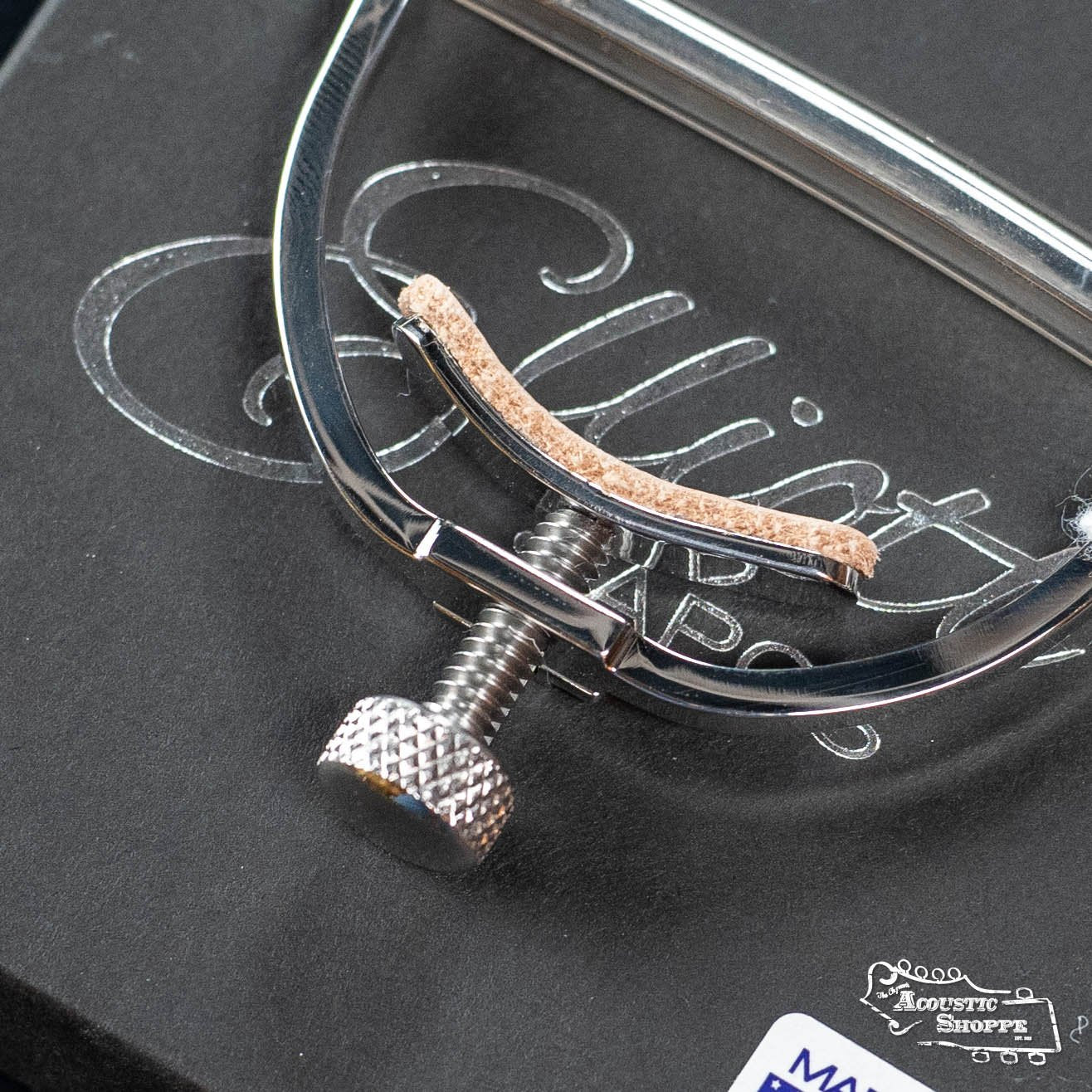 A close-up of the Elliott Capos G34E Push Button Elite Capo (1 3/4, 16 radius) with a cork pad, resting on a black box engraved with Elliott and featuring the Acoustic Shoppe logo—perfect for acoustic guitar enthusiasts.