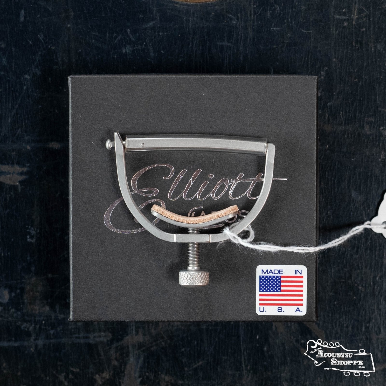 An Elliott G34E-S Elite Guitar Capo-Satin 1 3/4" with 16" Radius sits on a black Elliott Capos box bearing a "Made in U.S.A." sticker, American flag, and white tag, all on a dark textured surface.