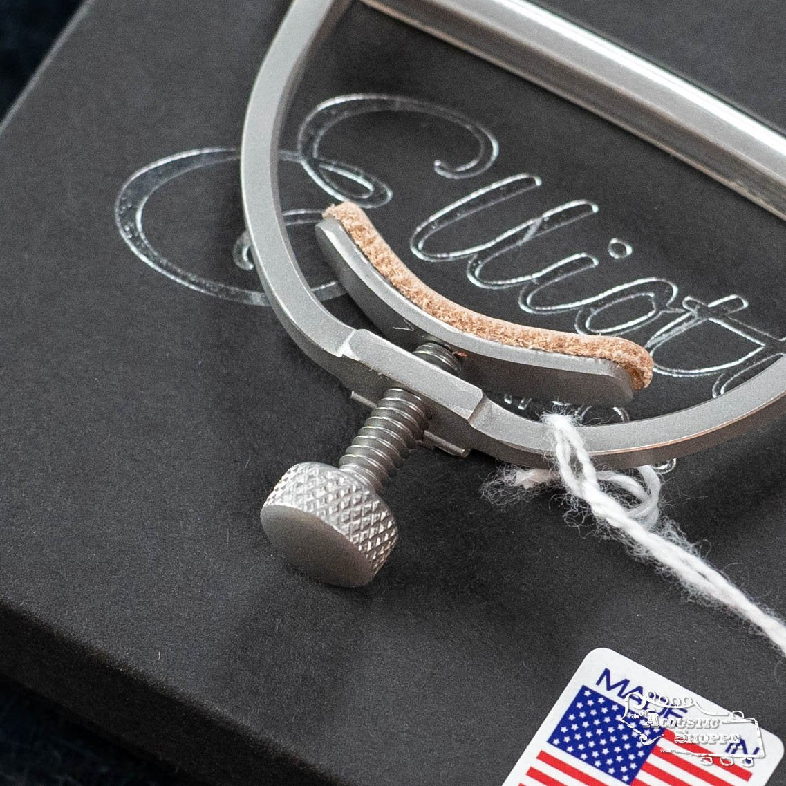 A close-up of the Elliott Capos G34E-S Elite Guitar Capo-Satin 1 3/4 w/ 14 Radius, featuring a textured adjustment screw, rests on a black box with cursive writing. A white string is attached and an Eastman dealer sticker appears in the lower right corner.