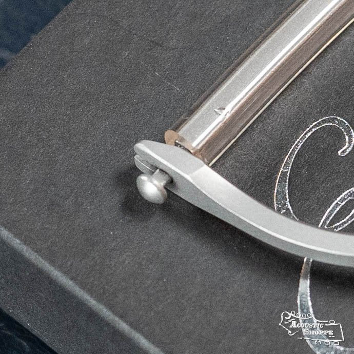 A close-up of the Elliott Capos G34E-S Elite Guitar Capo (Satin, 1 3/4 w/ 14 Radius) rests on a dark box, with partially visible decorative white text reading Acoustic Shoppe—a trusted Eastman dealer.