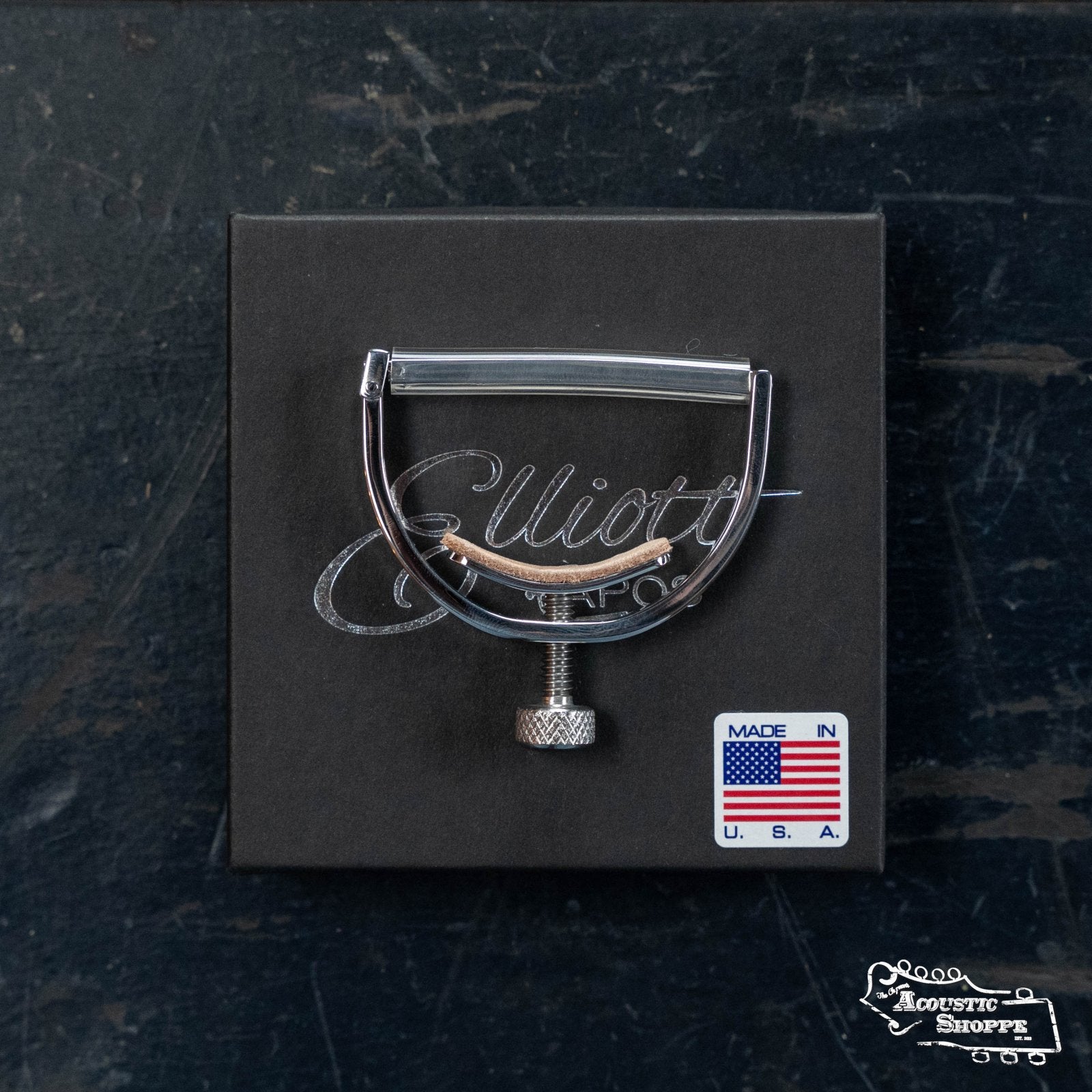 An Elliott G34M McKinney Guitar Capo 1 3/4" w/ 15" Radius by Elliott Capos is shown on a black box with the brand's script logo and a small "Made in U.S.A." flag sticker, all resting on a dark, textured surface.