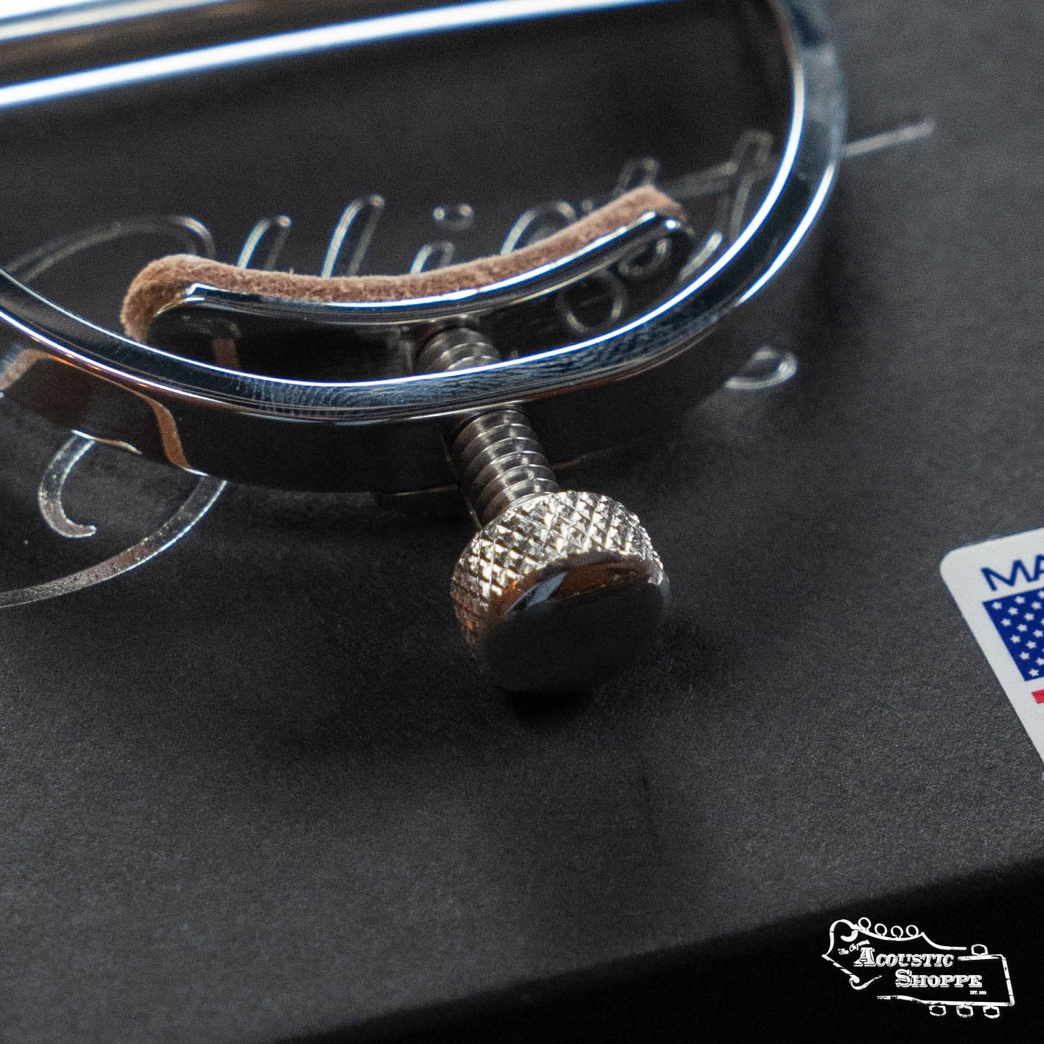 Close-up of the Elliott G34M McKinney Guitar Capo 1 3/4" w/ 15" Radius by Elliott Capos, featuring a knurled adjustment screw, resting on a black box with part of a logo and a Made in USA label visible.