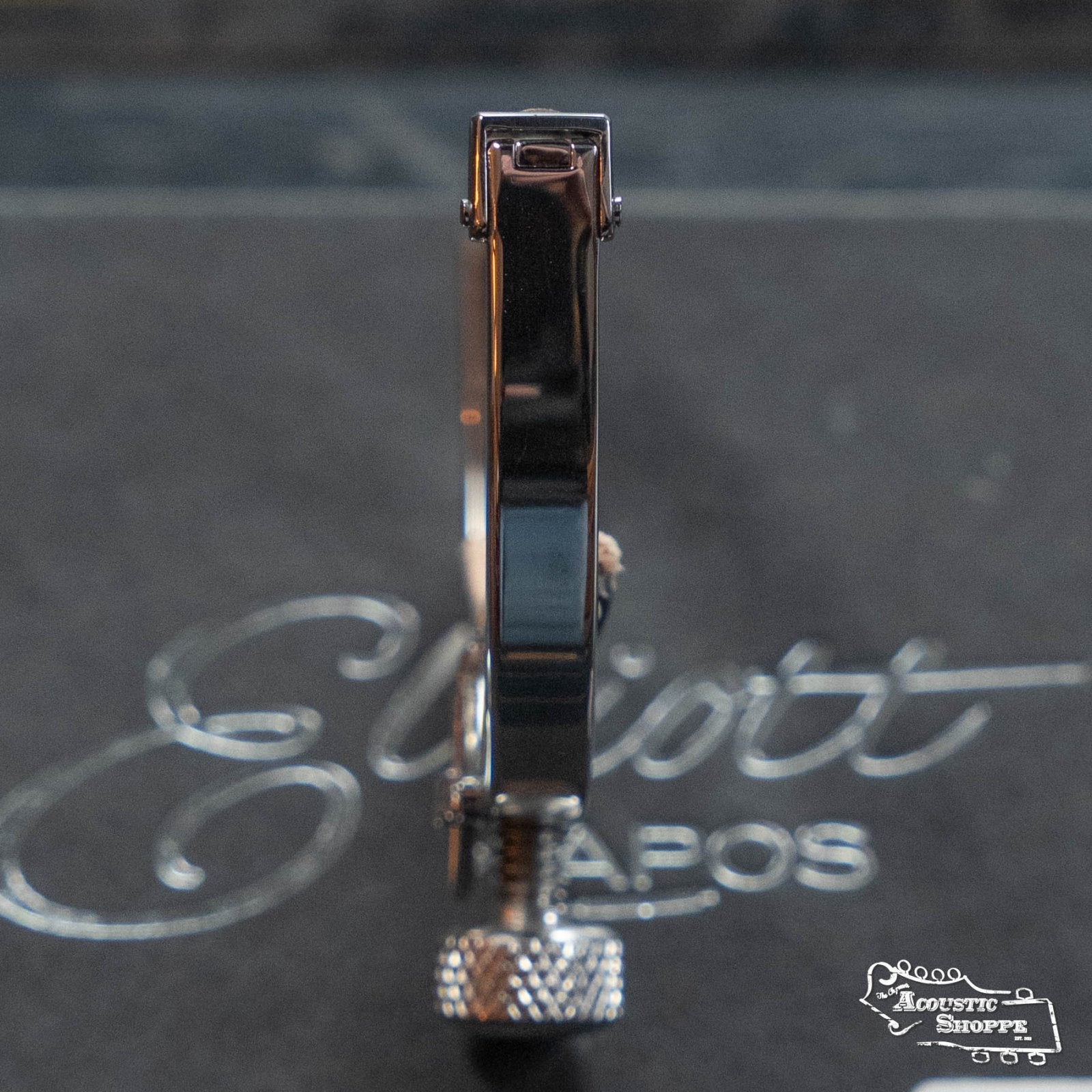 A close-up side view of the Elliott G34M McKinney Guitar Capo 1 3/4" w/ 15" Radius by Elliott Capos, upright on a surface with "Elliott Capos" and "Acoustic Shoppe" faintly visible in the background.