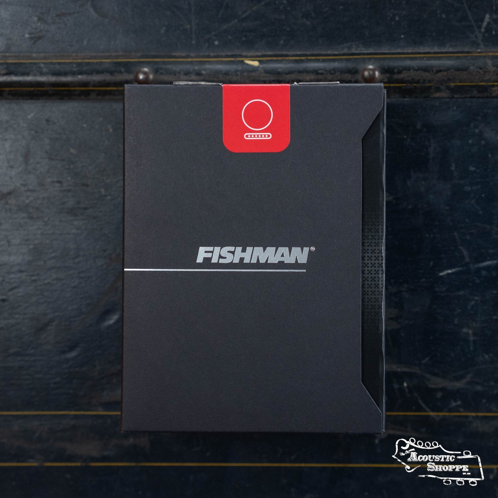 A black rectangular Fishman AG-Series Undersaddle Pickup box (PRO-AG0-094) for 6-string guitar, with a red label on top, rests on a dark wooden surface. The silver and white branding suggests quality from Fishman.