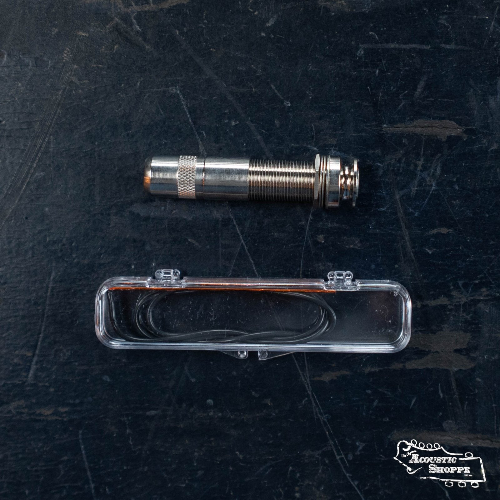 A Fishman AG-Series Undersaddle Pickup For 6-string guitar (PRO-AG0-094), including a metal input jack and clear plastic case with wires, rests on a dark textured surface. The Acoustic Shoppe logo appears in the bottom right corner.