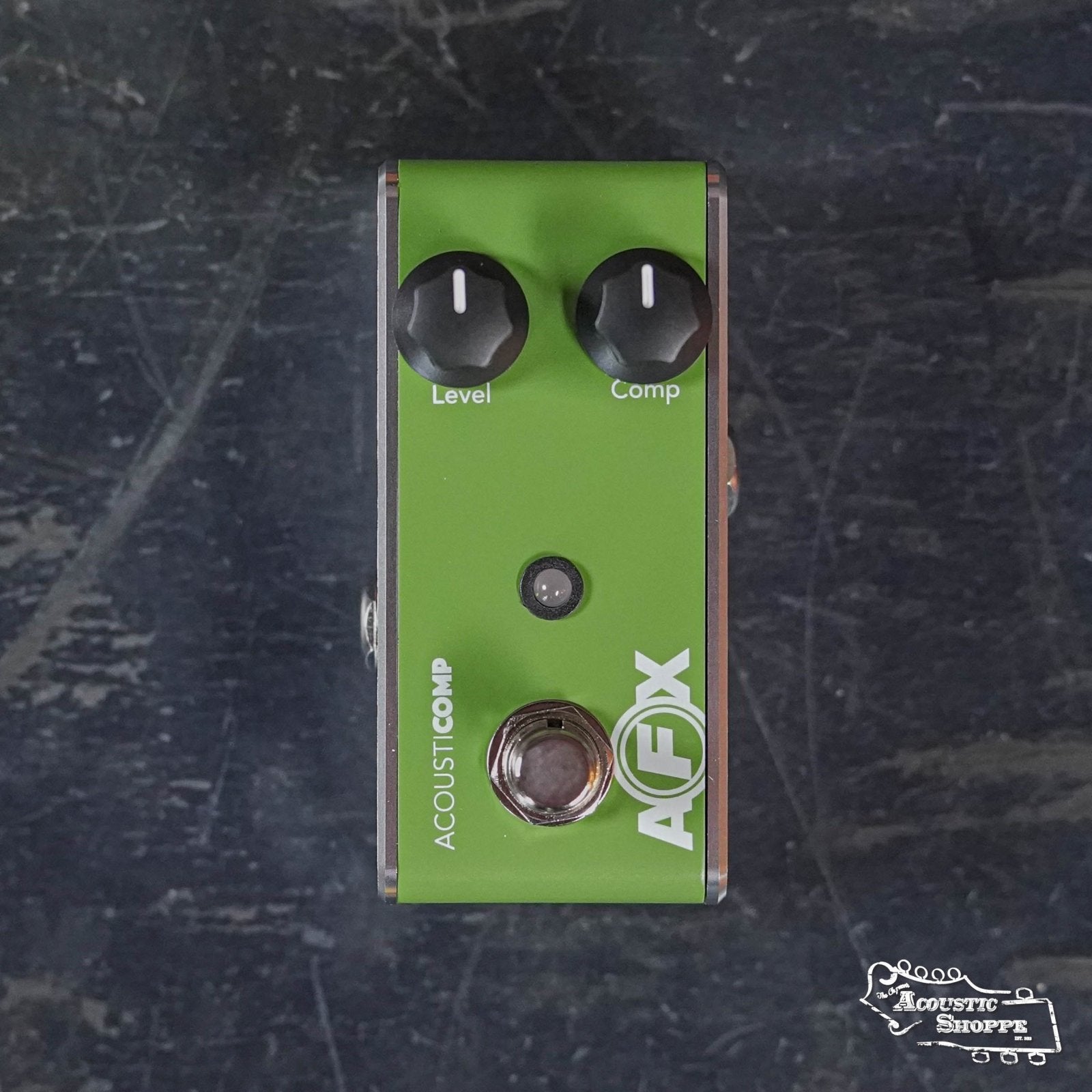 A green Fishman AFX AcousticComp Mini Compressor PRO-AFX-CM2 pedal with Level and Comp knobs sits on a dark, textured surface. The Acoustic Shoppe logo is in the bottom right, and it features true bypass for clean signal integrity.