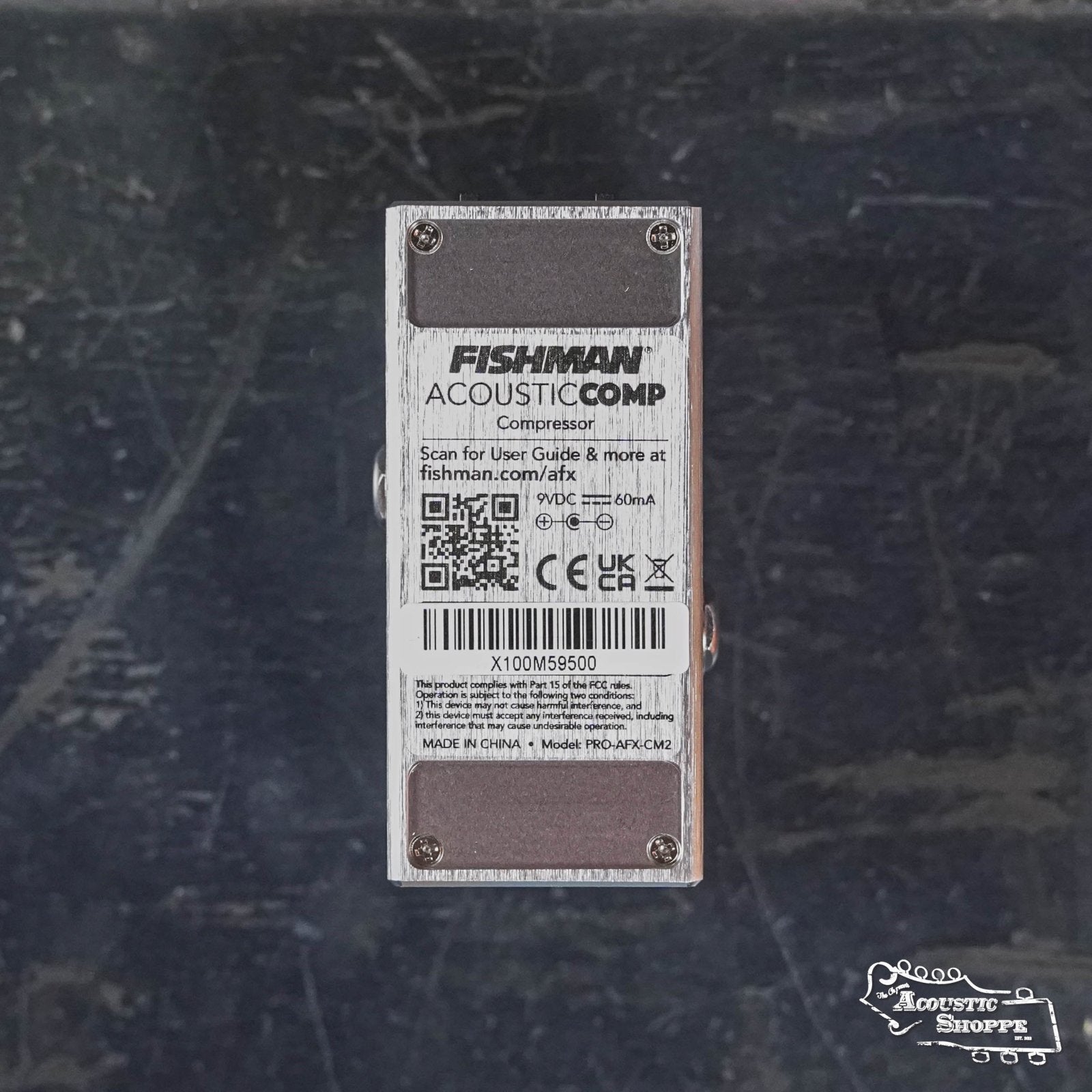 A Fishman AFX AcousticComp Mini Compressor PRO-AFX-CM2 pedal viewed from the bottom, displaying a label with text, QR code, barcode, and product details on a dark textured background—featuring true bypass for signal clarity.