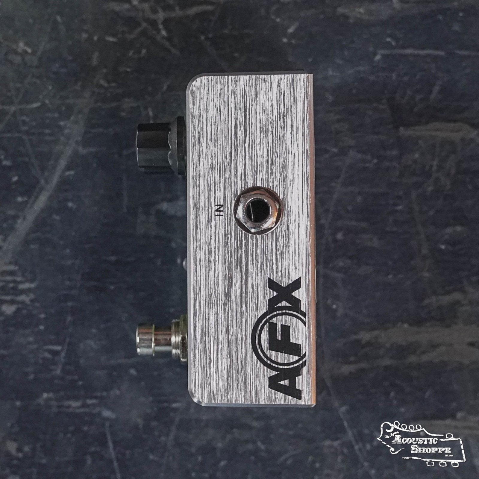 A Fishman AFX AcousticComp Mini Compressor PRO-AFX-CM2 pedal, with input jack, control knob, and true bypass footswitch, rests on a dark textured surface. The Acoustic Shoppe logo is visible in the bottom right corner.