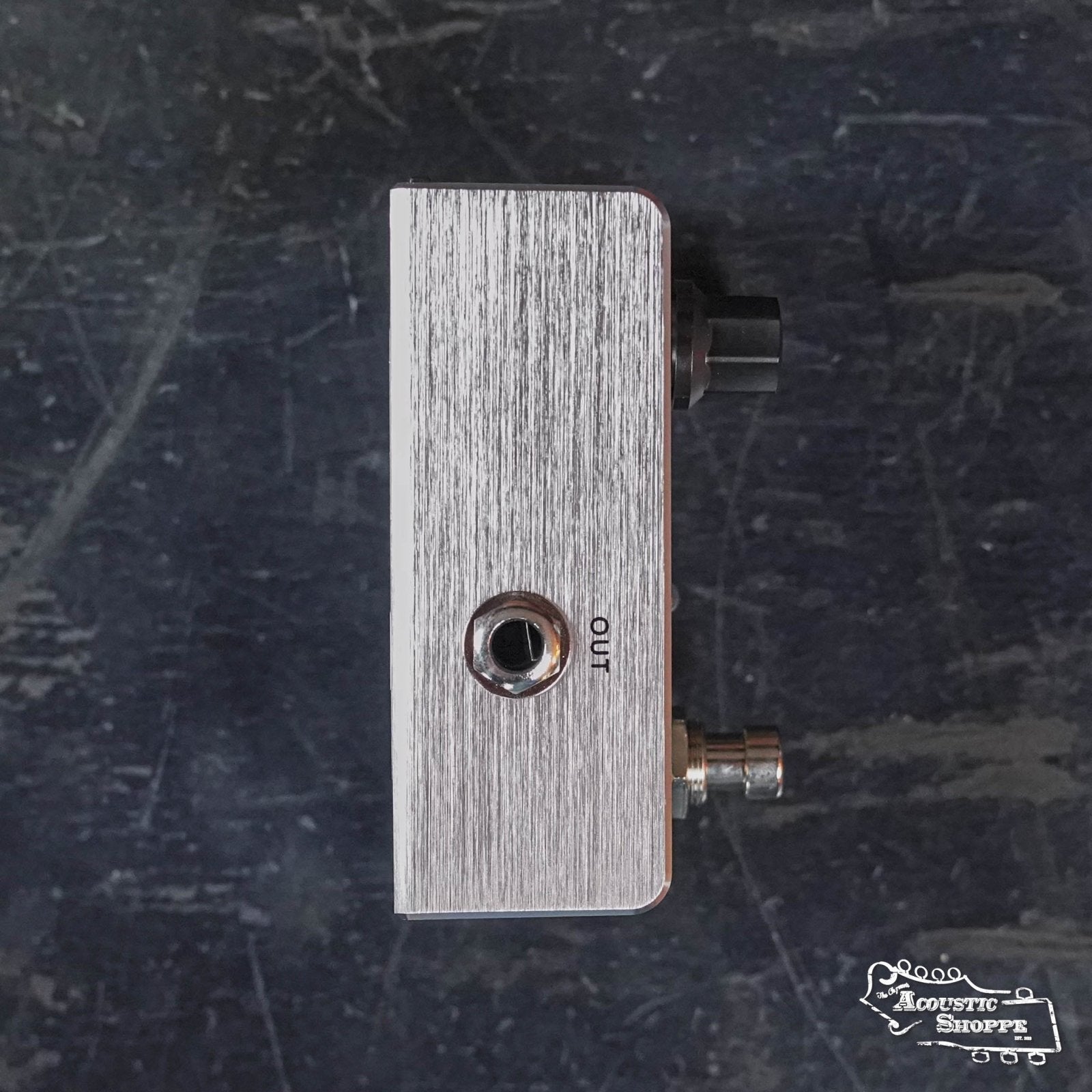 A Fishman AFX AcousticComp Mini Compressor PRO-AFX-CM2 pedal in silver is shown from the side, displaying the OUT jack, black knob, and footswitch on a dark textured surface with The Acoustic Shoppe logo in white.