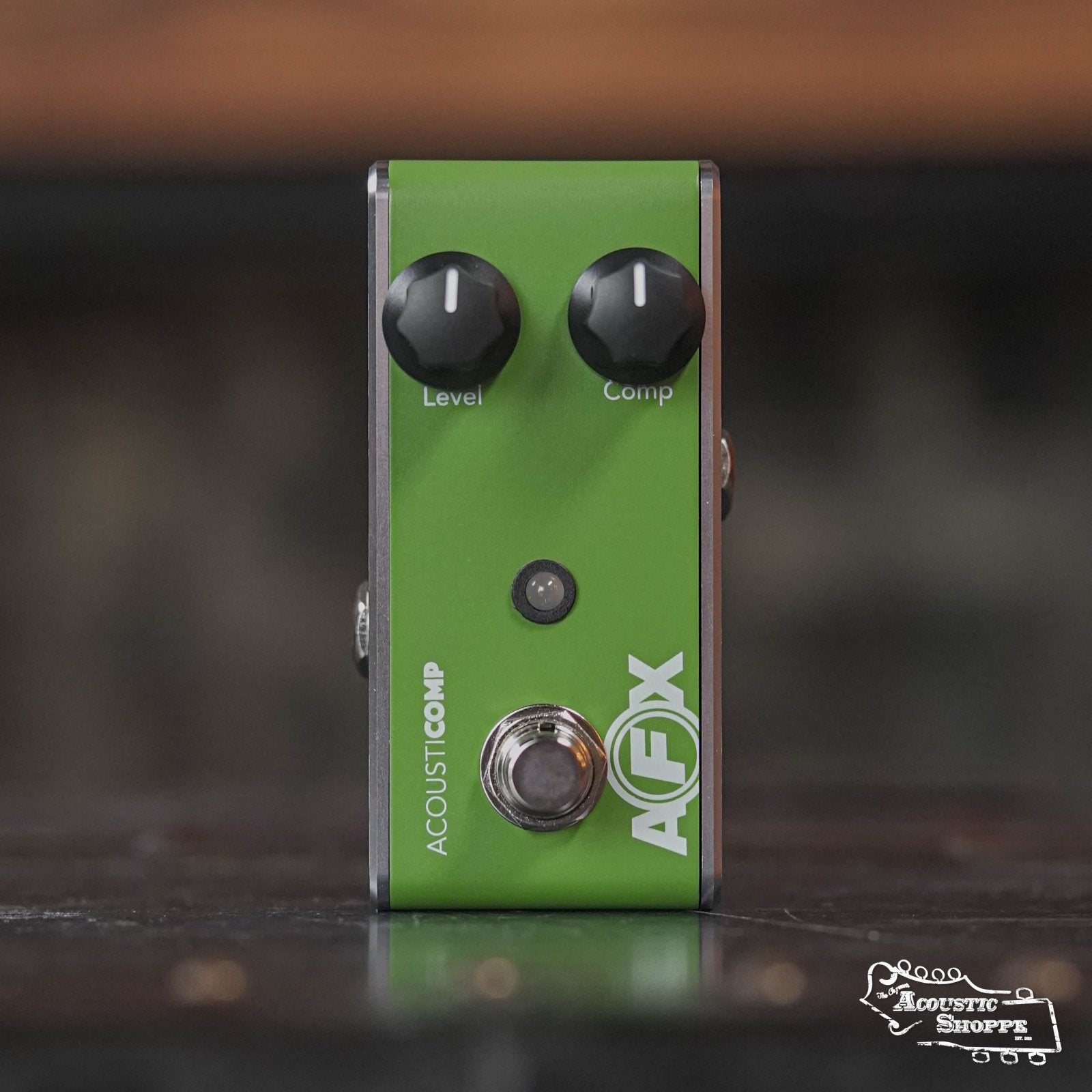 A green Fishman AFX AcousticComp Mini Compressor PRO-AFX-CM2 stands upright on a dark surface with two black knobs labeled "Level" and "Comp." The Acoustic Shoppe logo is visible in the bottom right corner.