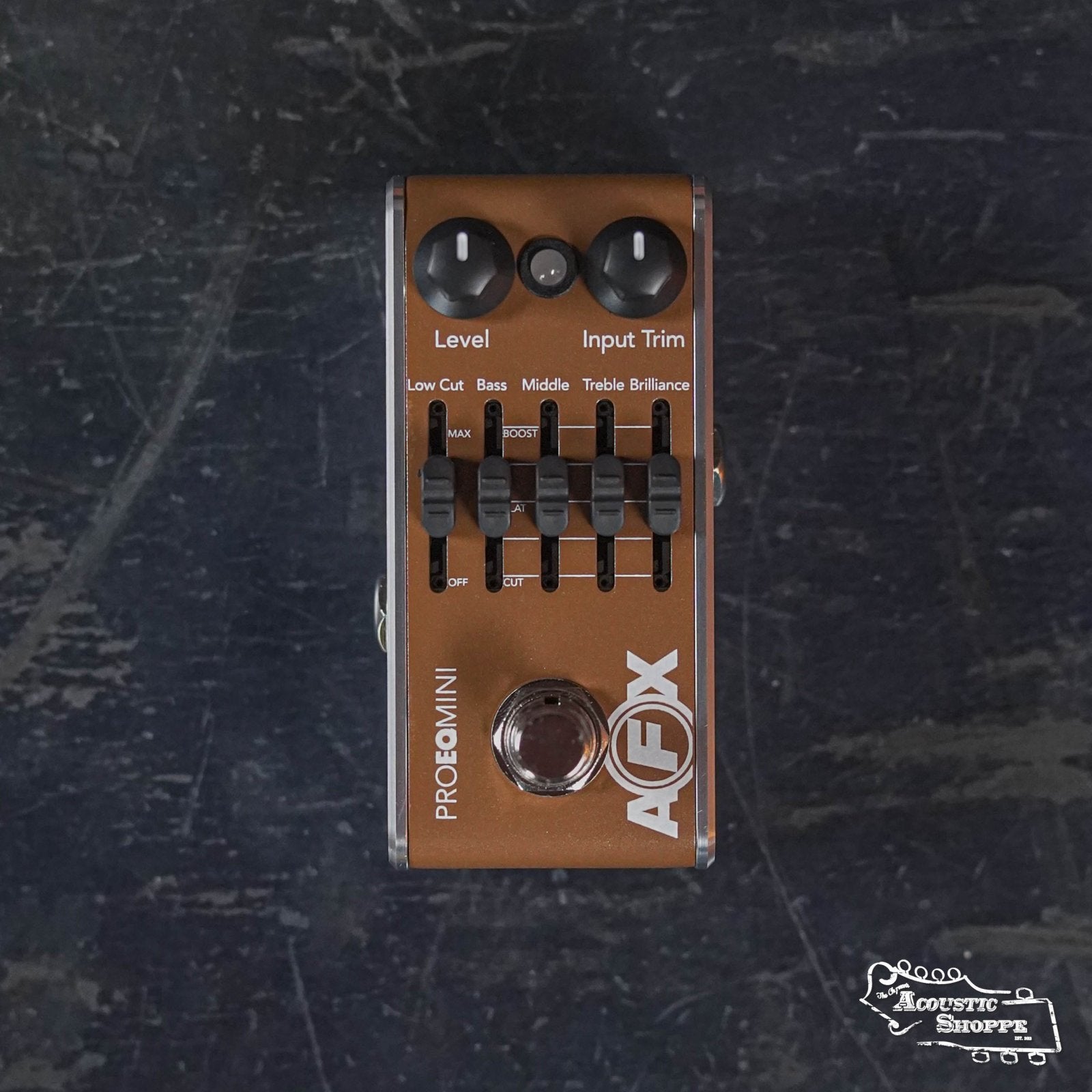 A Fishman AFX Pro EQ Mini Acoustic Preamp & EQ PRO-AFX-EQ2 pedal with five sliders and two knobs lies on a dark textured surface, offering level, input trim, EQ band controls, and a low cut switch.