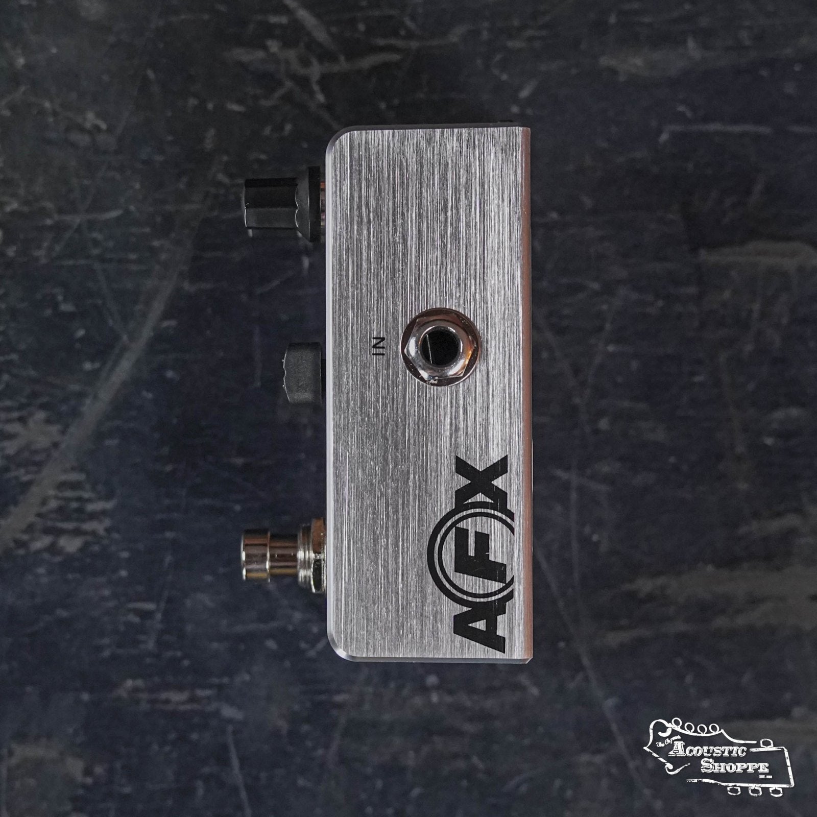 Side view of a metallic Fishman AFX Pro EQ Mini Acoustic Preamp & EQ PRO-AFX-EQ2 pedal, showing input/output jacks, on a dark scratched surface. The Acoustic Shoppe logo is in the bottom right corner.