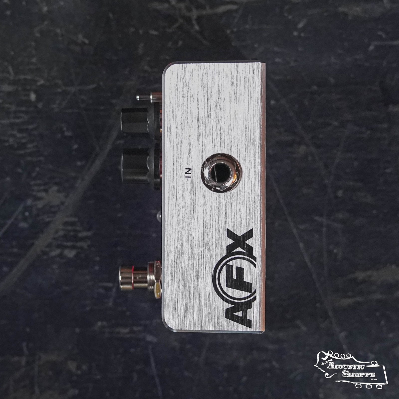 A Fishman AFX AcoustiVerb Mini Reverb Pedal PRO-AFX-RV2 is seen from the side, highlighting the IN input jack and black knobs. The AFX name and Acoustic Shoppe logo are visible on a dark textured surface—ideal for acoustic guitar reverb effects.