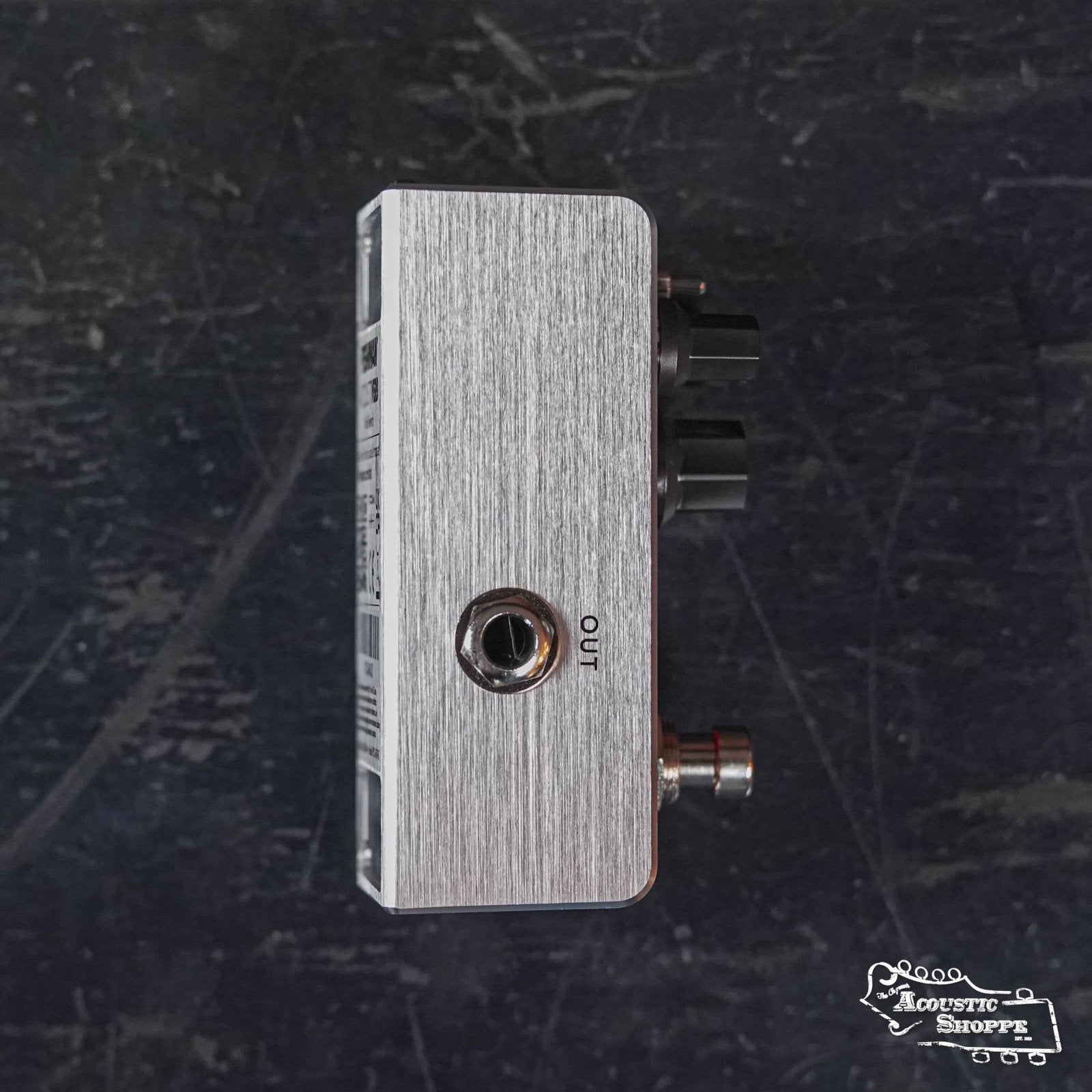 Side view of the Fishman AFX AcoustiVerb Mini Reverb Pedal PRO-AFX-RV2 in silver, featuring an OUT jack and two black knobs, on a worn dark surface. The Acoustic Shoppe logo is at bottom right. Ideal for acoustic guitar reverb effects.