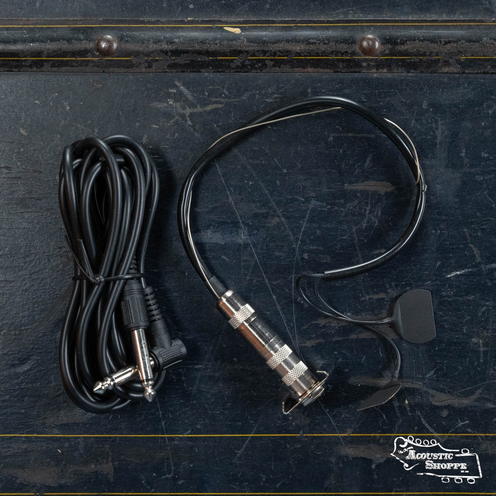 The Fishman BP-100 Upright Bass Pickup (PRO-BP1-00B) by Fishman, featuring a long black cable, quarter-inch plug, and small microphone attachment, is displayed on a dark textured surface with The Acoustic Shoppe logo in the bottom right corner.