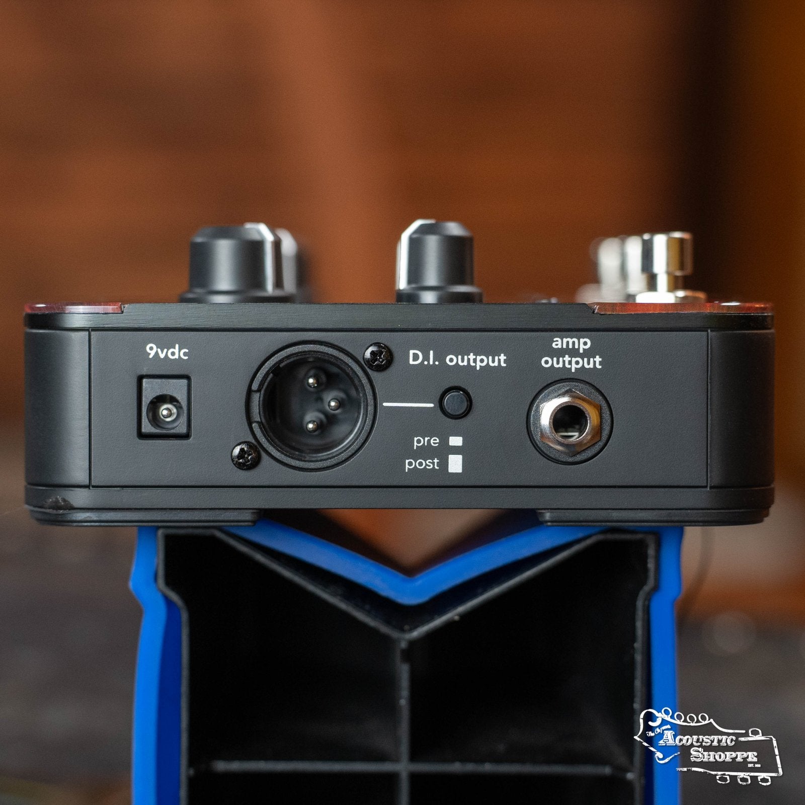 Close-up of the Fishman ToneDEQ Preamp EQ (PRO-DEQ-AFX) by Fishman, featuring a 9V DC input, D.I. output with pre/post switch, and amp output. Perfect for acoustic musicians, it rests on a blue stand with a blurred brown background.
