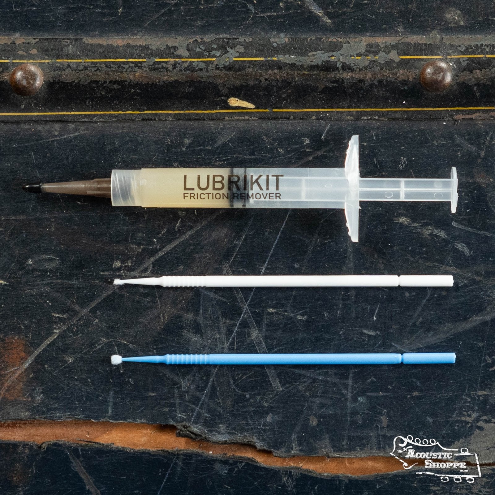 A DAddario PW-LBK-01 LubriKit Friction Remover syringe and two thin applicator brushes—one white, one blue—lie on a dark surface, perfect for lubricating guitar nuts to improve tuning stability.