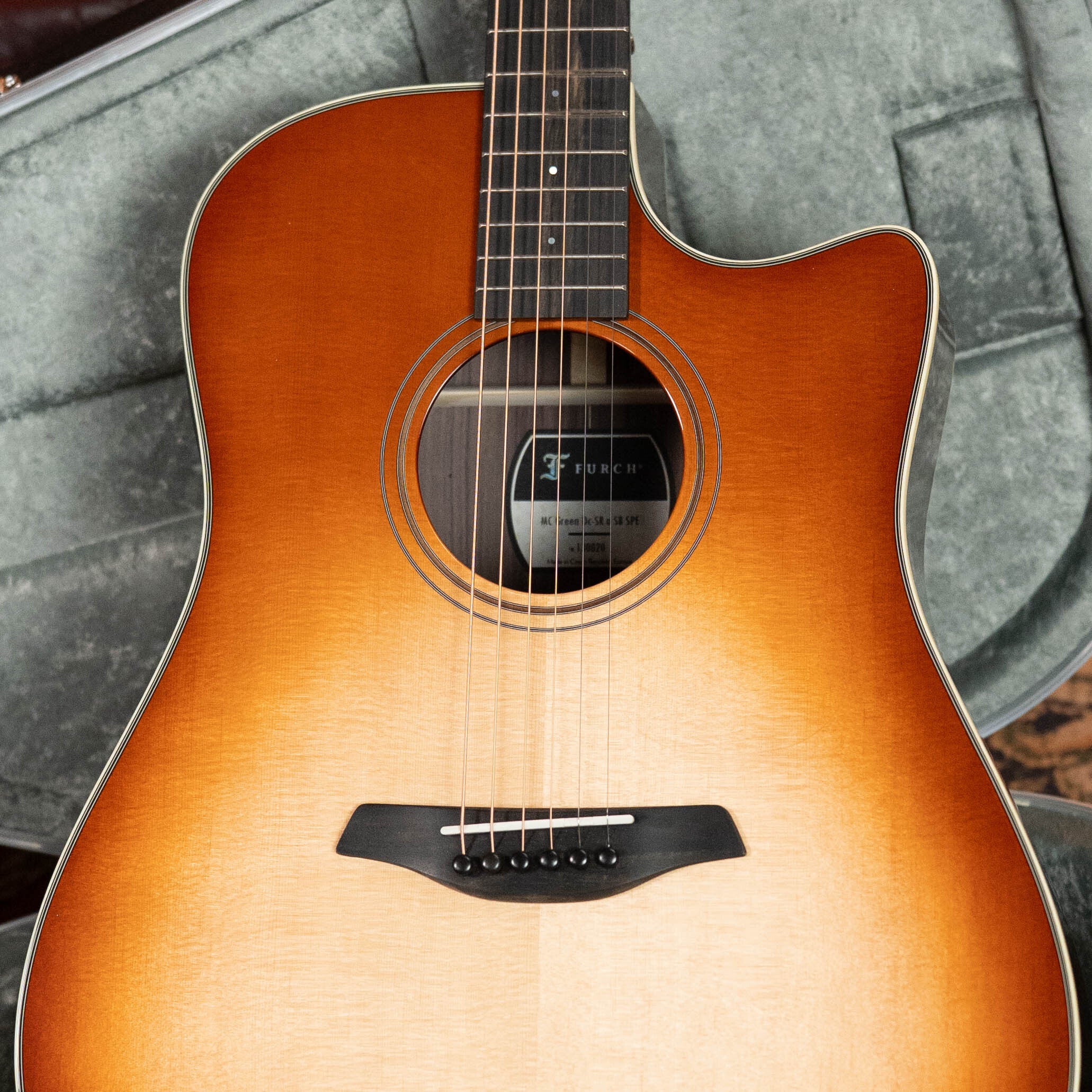 A close-up of the Furch Masters Choice Green SB Dc-SR AA Sitka/AA Indian Rosewood Dreadnought Cutaway Sunburst Guitar with LR Baggs Pickup, showing its body, strings, and “Furch” label inside a gray padded case.