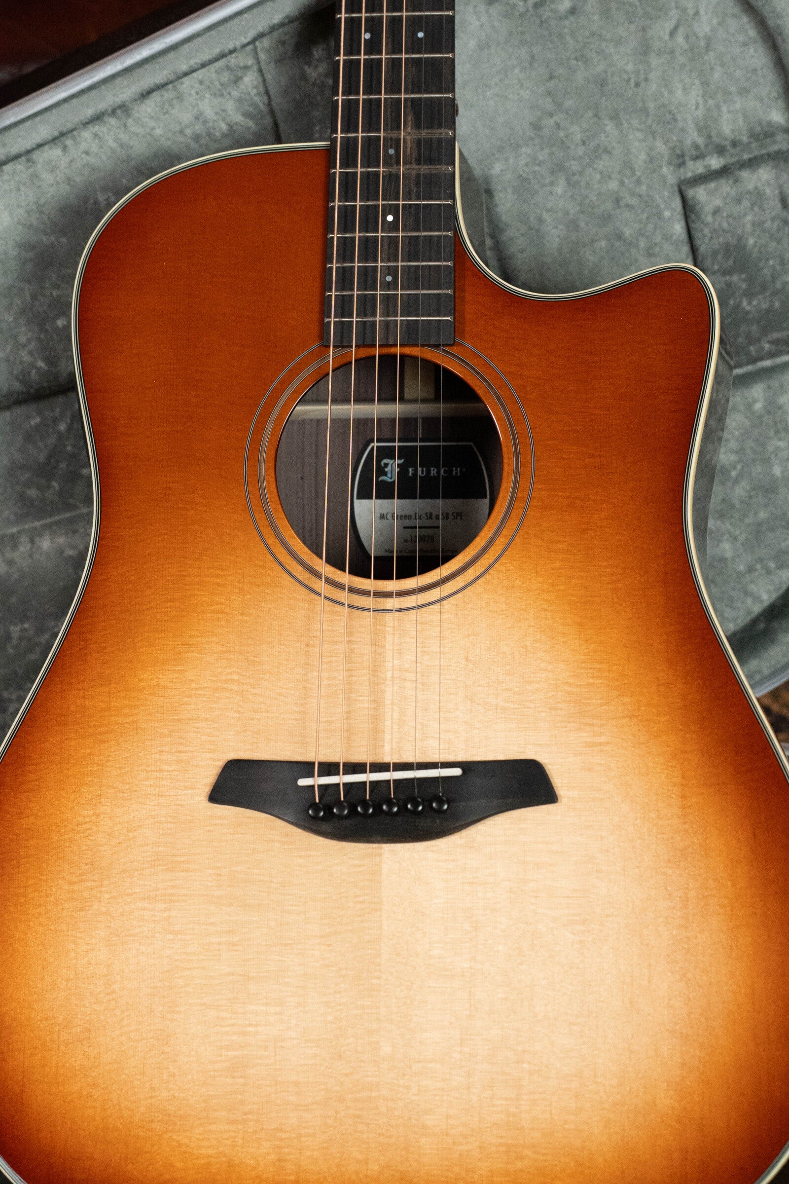 A close-up of the Furch Masters Choice Green SB Dc-SR AA Sitka/AA Indian Rosewood dreadnought cutaway guitar in sunburst finish, showing its Sitka spruce top, strings, sound hole, and part of the fretboard on a gray textured surface.