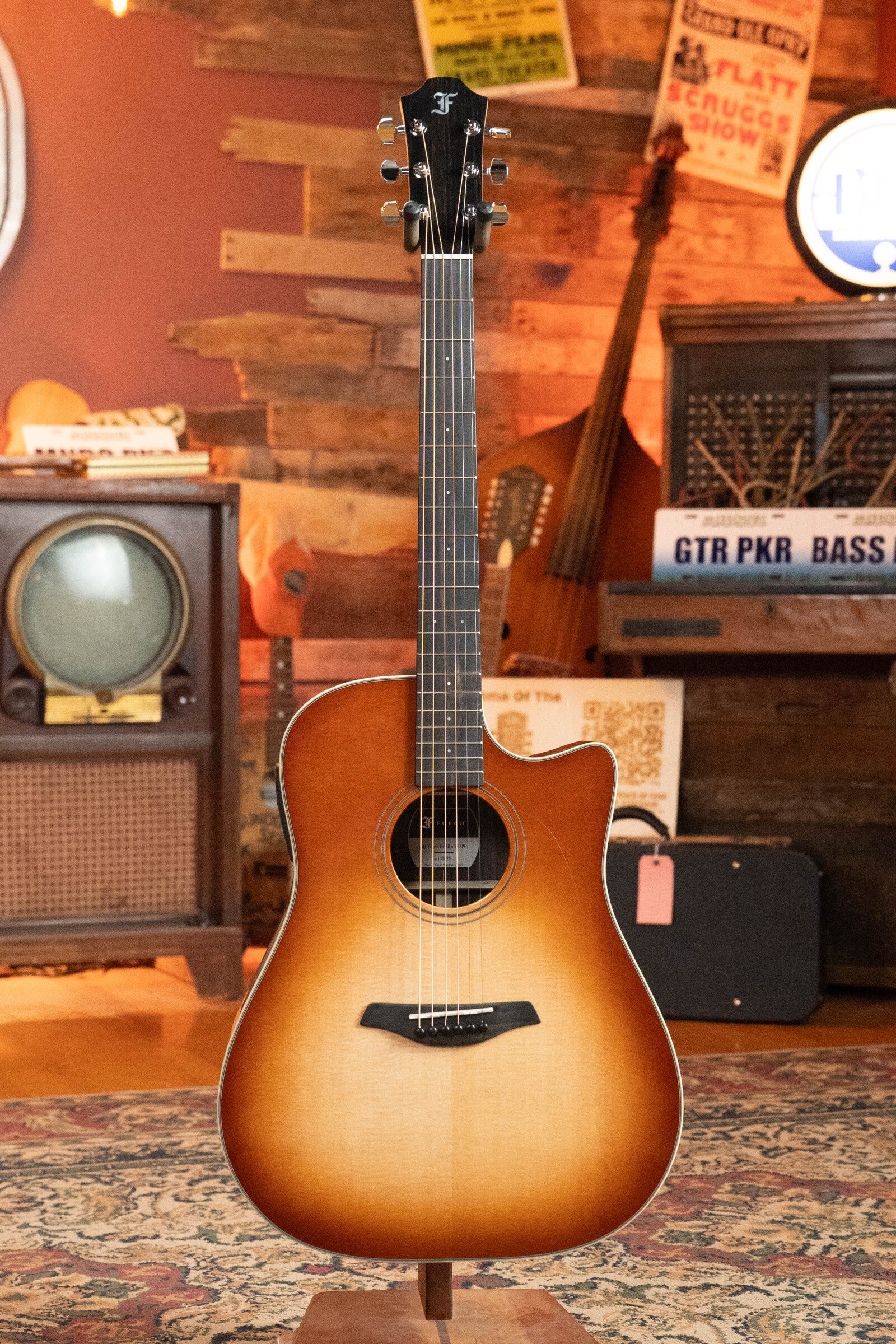 A Furch Masters Choice Green SB Dc-SR AA Sitka/AA Indian Rosewood Dreadnought Cutaway Sunburst Guitar w/LR Baggs Pickup #0020 is displayed on a stand in a cozy, vintage-style room with wood paneling, retro TV, and instruments.