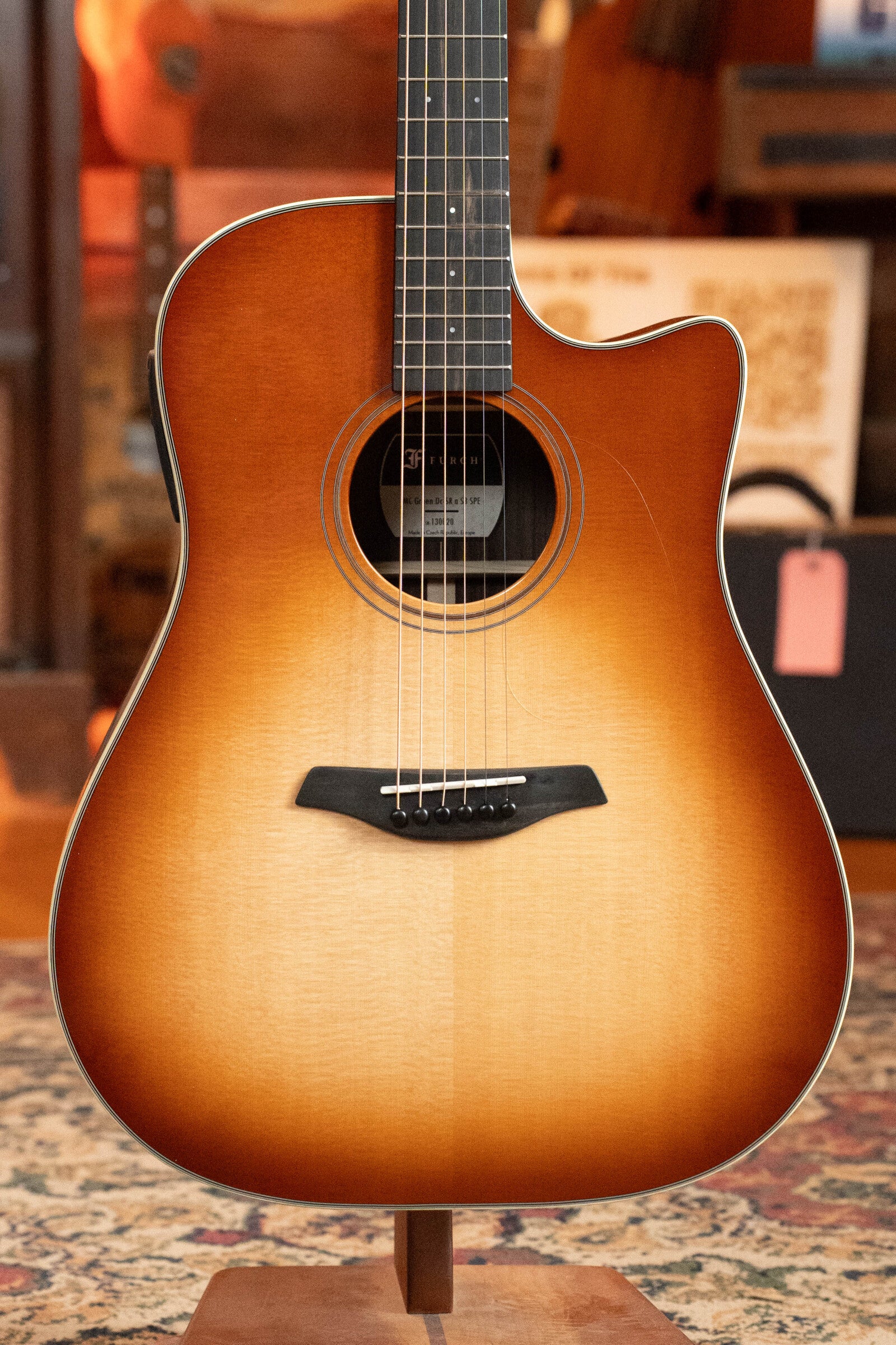 A Furch Masters Choice Green SB Dc-SR AA Sitka/AA Indian Rosewood Dreadnought Cutaway Sunburst Guitar w/LR Baggs Pickup is displayed on a stand, highlighting its Sitka spruce top, rich wood grain, and sunburst finish under warm lighting.