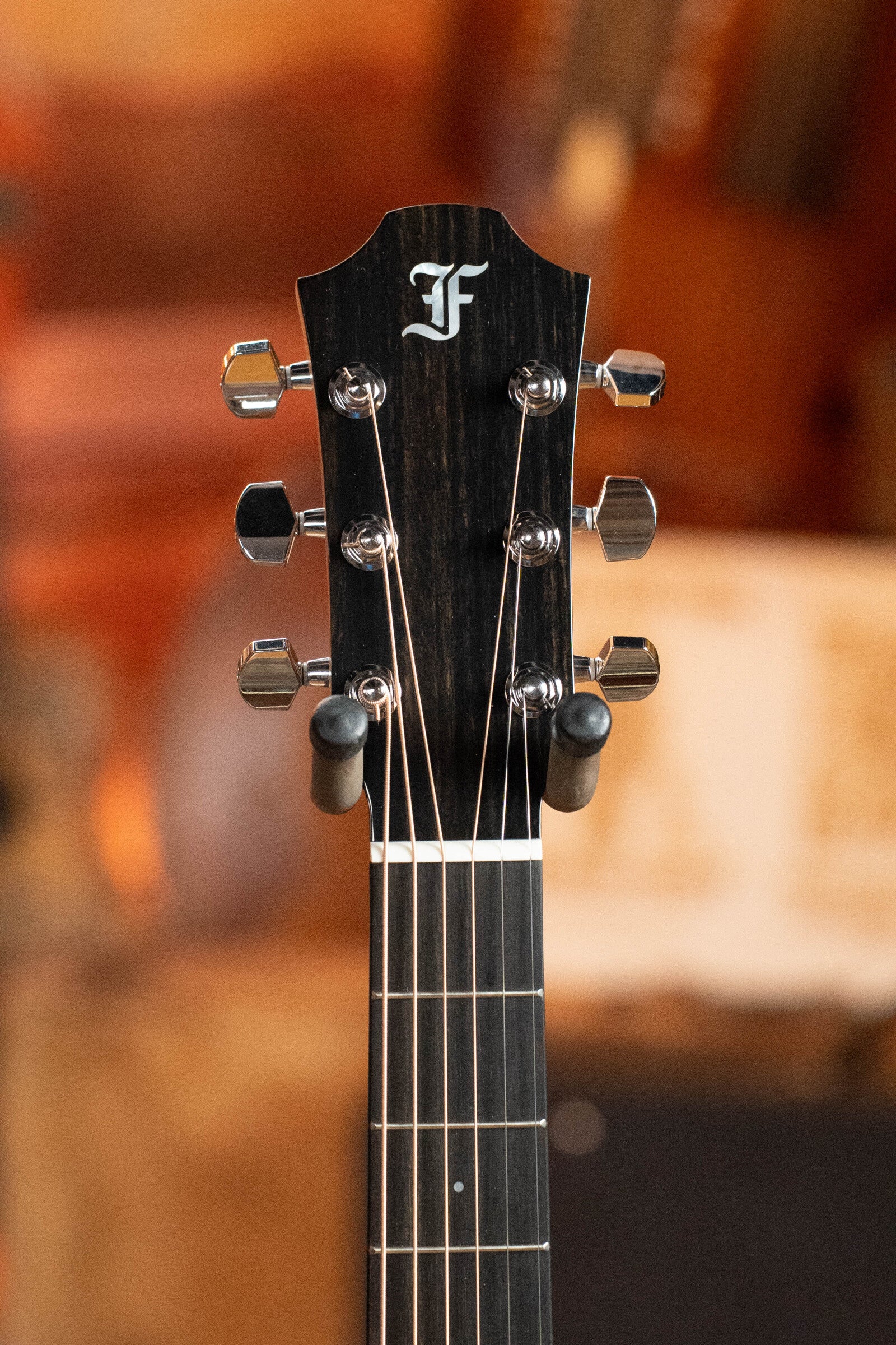 Close-up of a Furch Masters Choice Green SB Dc-SR AA Sitka/AA Indian Rosewood Dreadnought Cutaway Sunburst Guitar #0020 headstock with six tuning pegs and F logo, focusing on the strings and hardware against a warm, softly blurred backdrop.