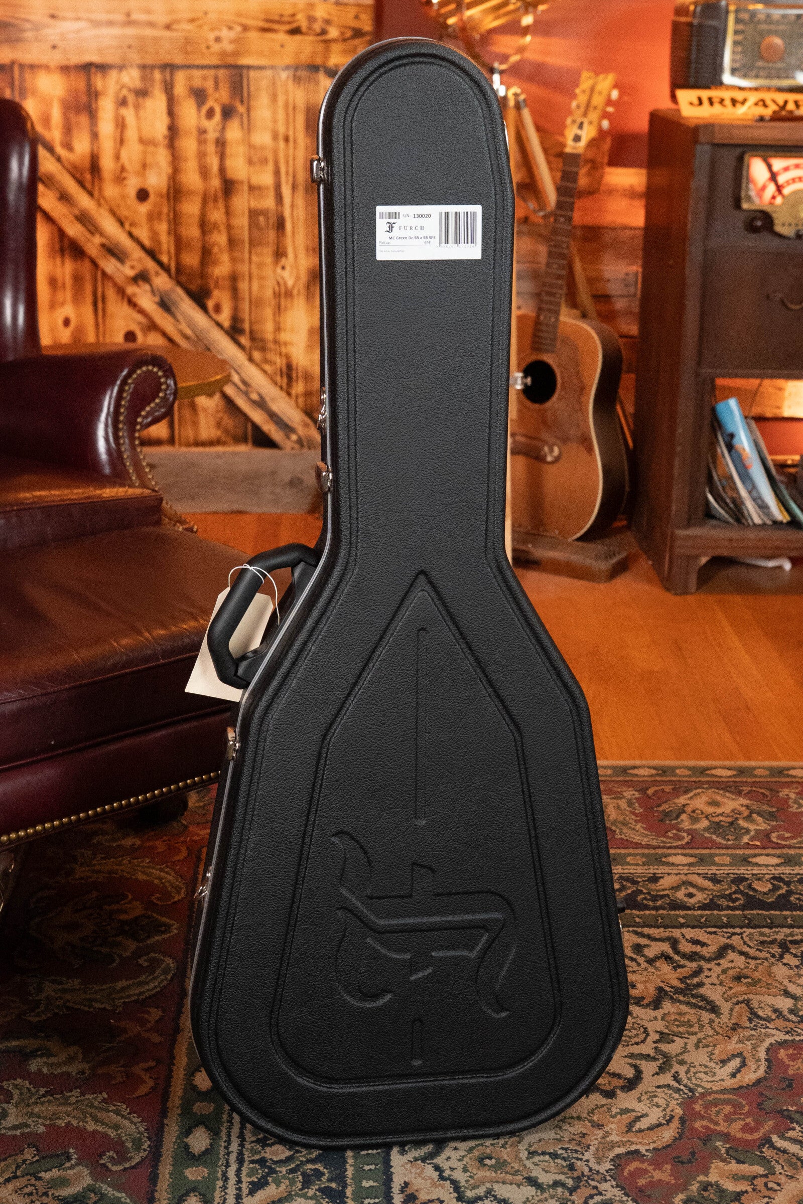 A black hard guitar case with a molded design stands upright on a patterned rug, ready to protect your Furch Masters Choice Green SB Dc-SR AA Sitka/AA Indian Rosewood Dreadnought Cutaway Sunburst Guitar.