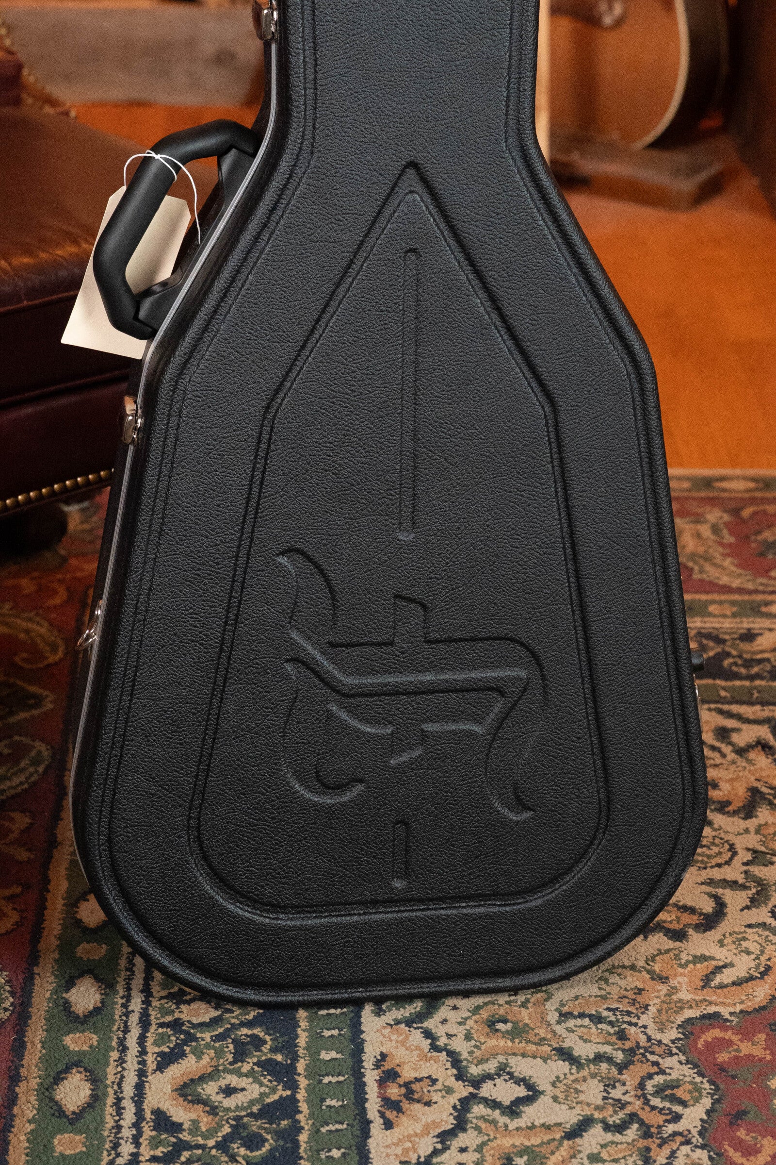 A black guitar case with an embossed logo stands on a patterned rug by a brown leather chair. Behind it, a Furch Masters Choice Green SB Dc-SR AA Sitka/AA Indian Rosewood Dreadnought Cutaway Sunburst Guitar w/LR Baggs Pickup is partially visible.