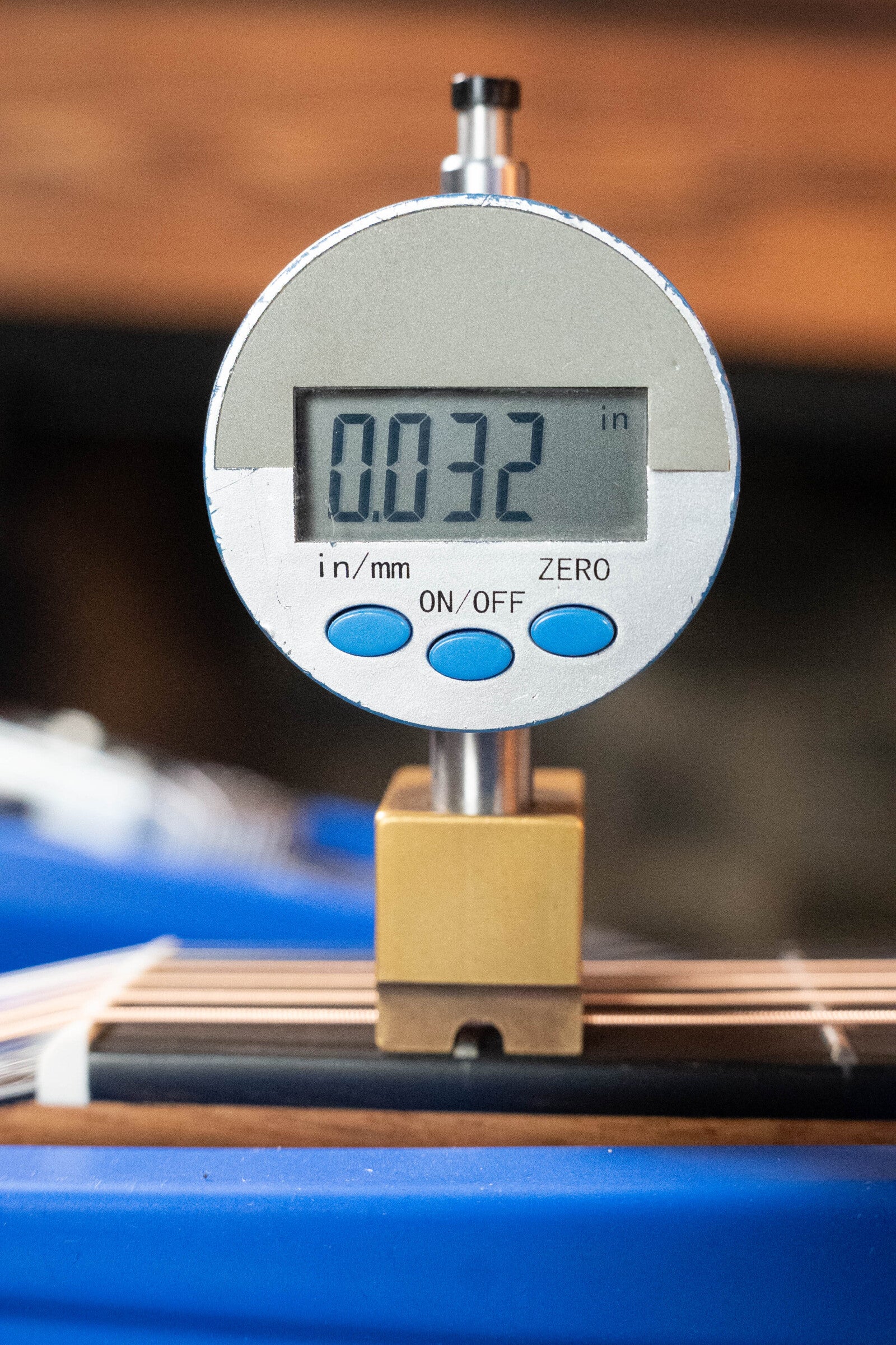 A digital gauge displays 0.032 inches while mounted on a stand, with blue and white objects blurred in the background—possibly tools for working on a Furch Masters Choice Green SB Dc-SR AA Sitka/AA Indian Rosewood Dreadnought Cutaway Sunburst Guitar.