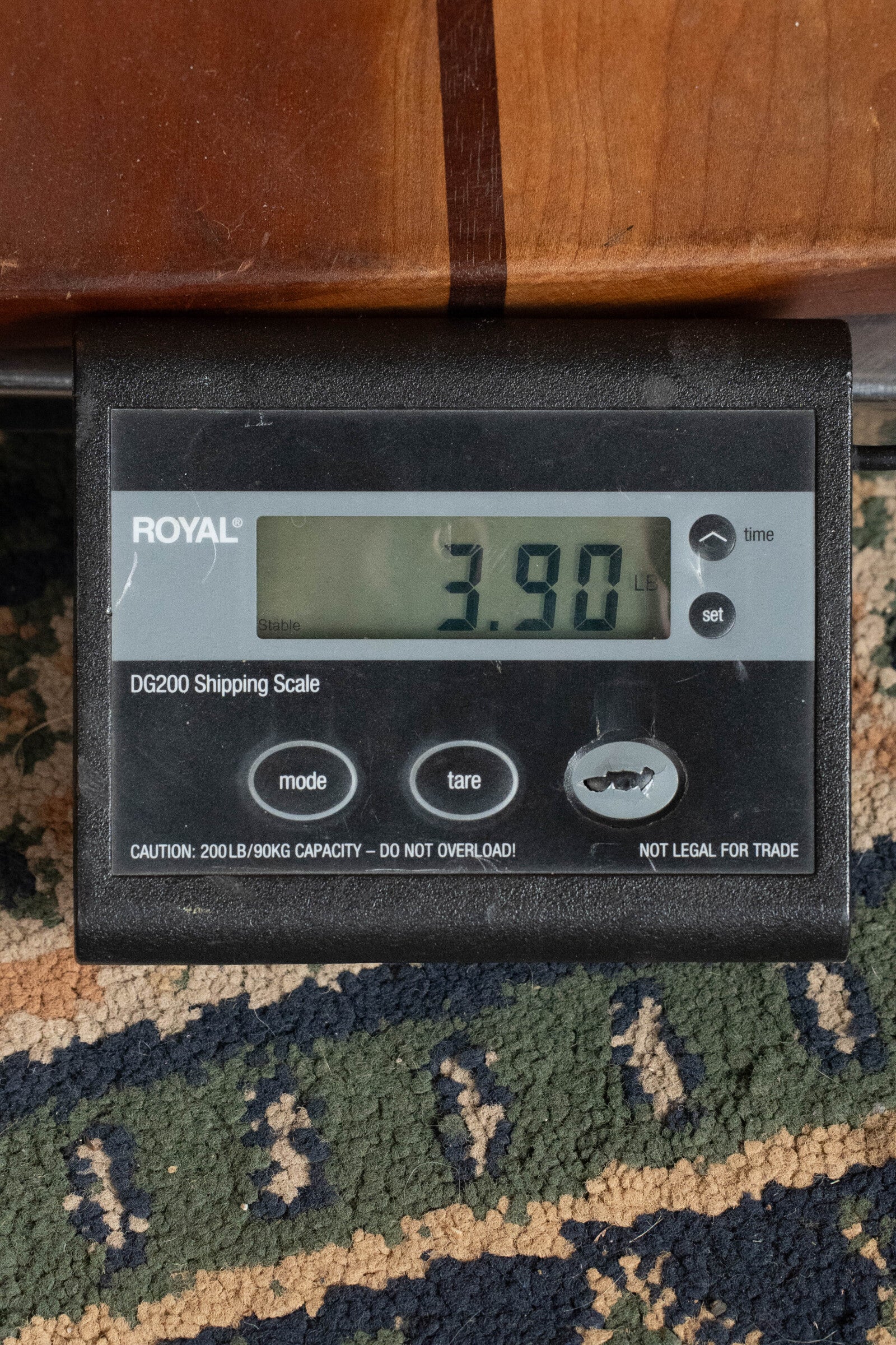 A Furch Pioneer-ER VTC Engelmann Spruce/Indian Rosewood Cutaway Travel Guitar rests near a digital shipping scale marked 3.90 lbs on patterned carpet, partially under wood. The scene suggests music with Furch branding visible on the scale.