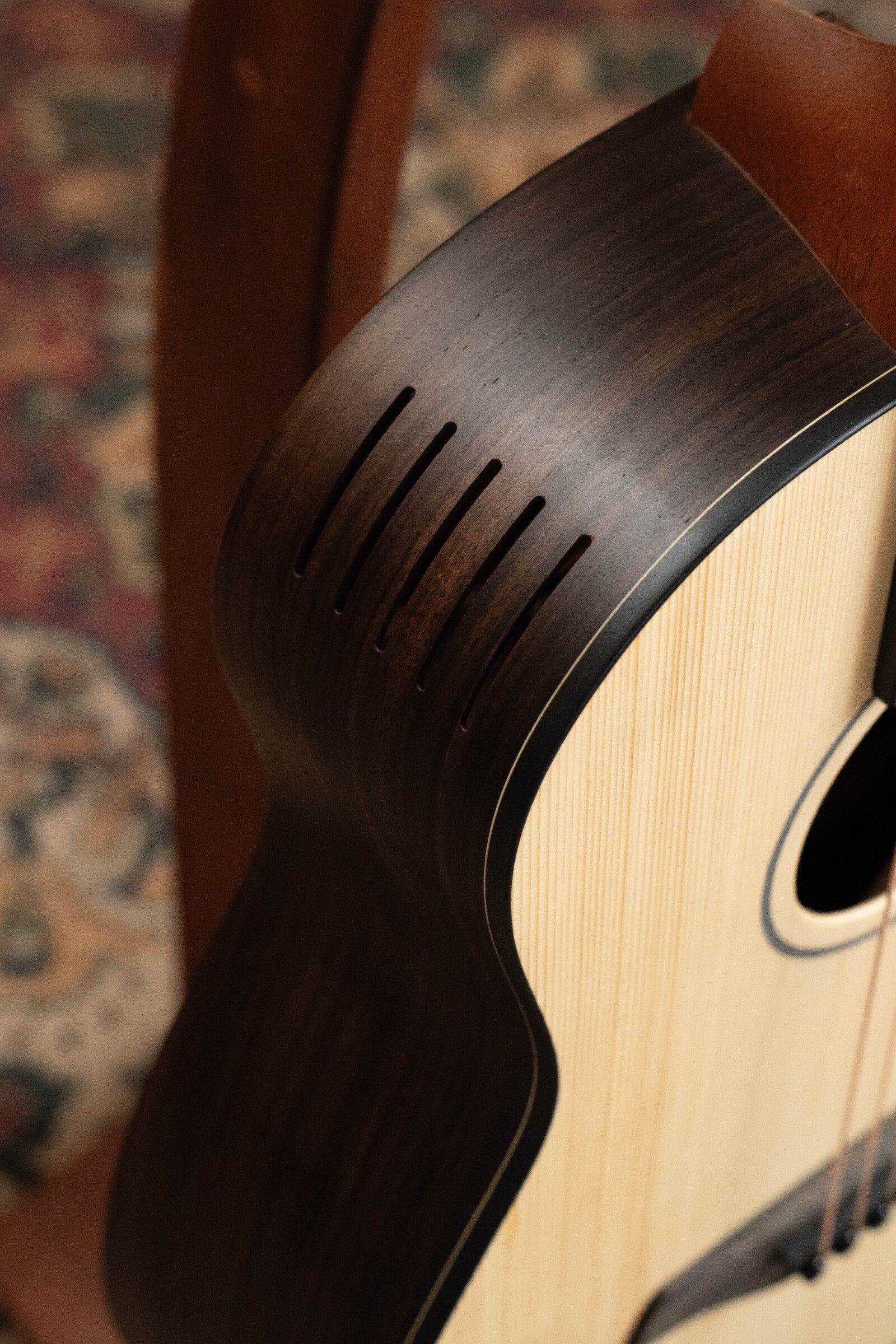 Close-up of the Furch Pioneer-ER VTC Cutaway Travel Guitar, showing its Indian Rosewood side, Engelmann Spruce top, and soundport boosters. A patterned rug and brown chair are blurred in the background.