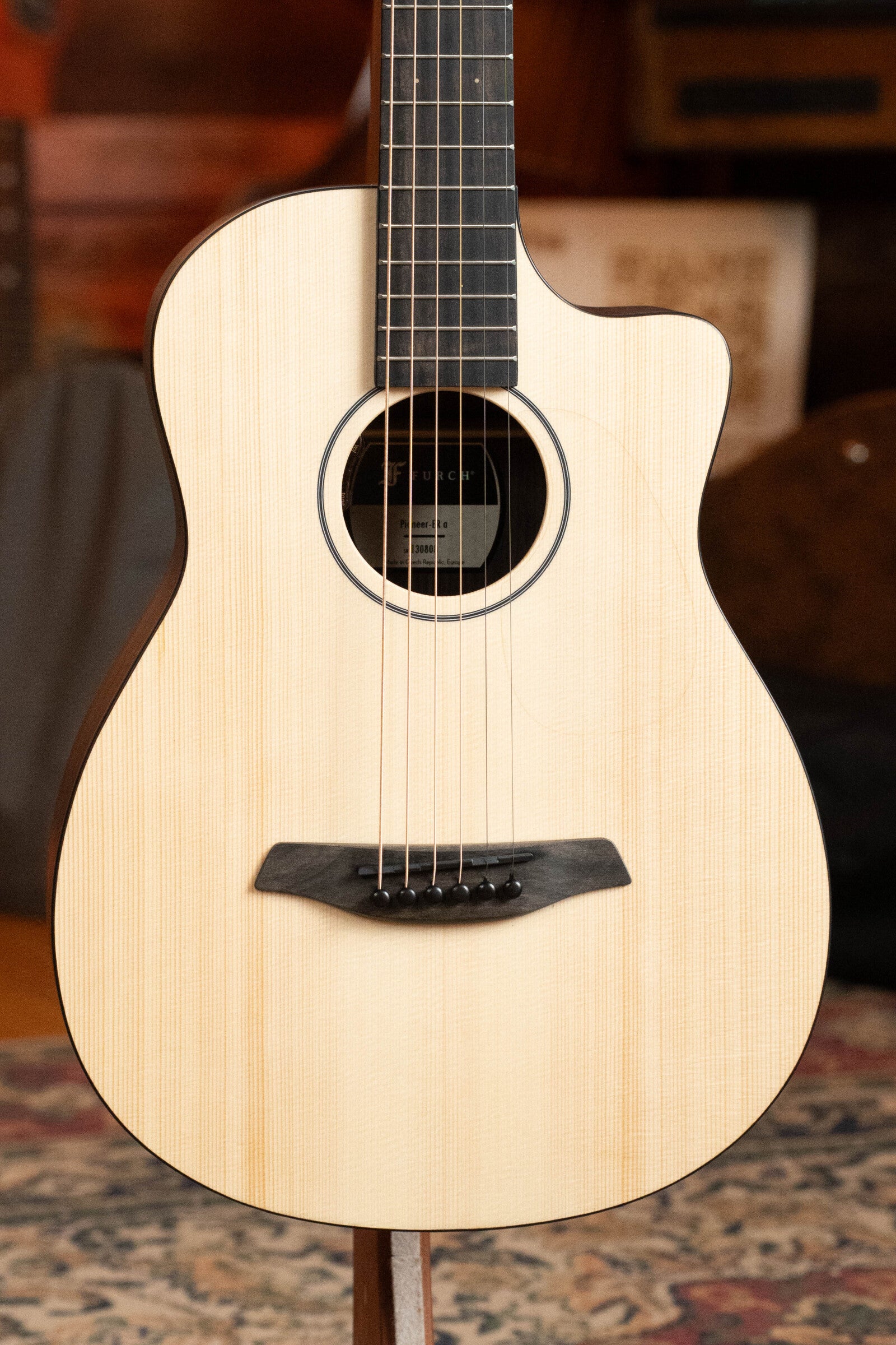 A close-up of the Furch Pioneer-ER VTC Travel Guitar, featuring an Engelmann Spruce top, Indian Rosewood back and sides, cutaway design, and VTC pickup, standing upright indoors on a patterned rug.