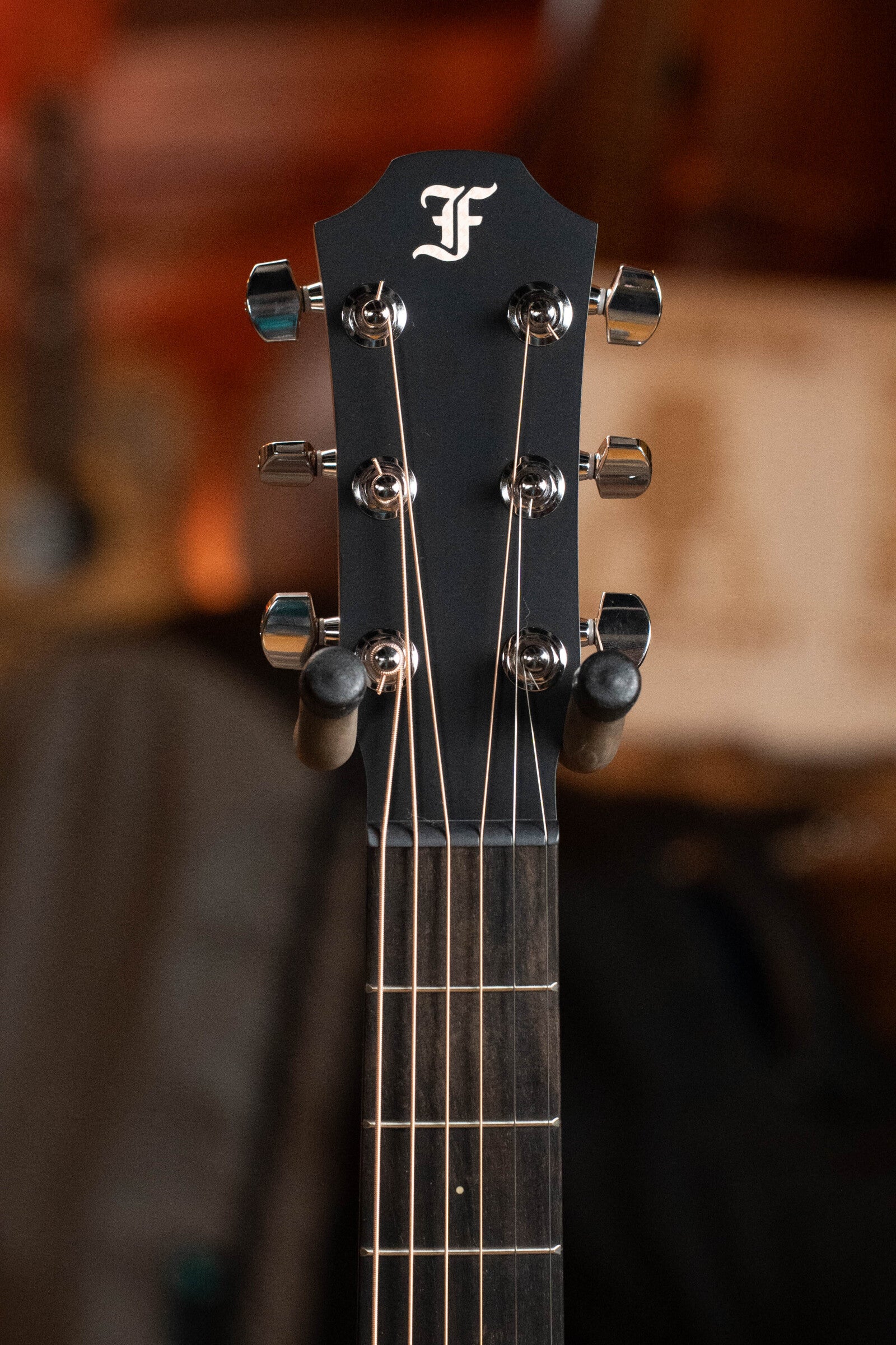 Close-up of the Furch Pioneer-ER VTC Travel Guitar headstock in black with six tuning pegs, F logo, and visible fretboard. Warm brown background is softly blurred for a refined effect.