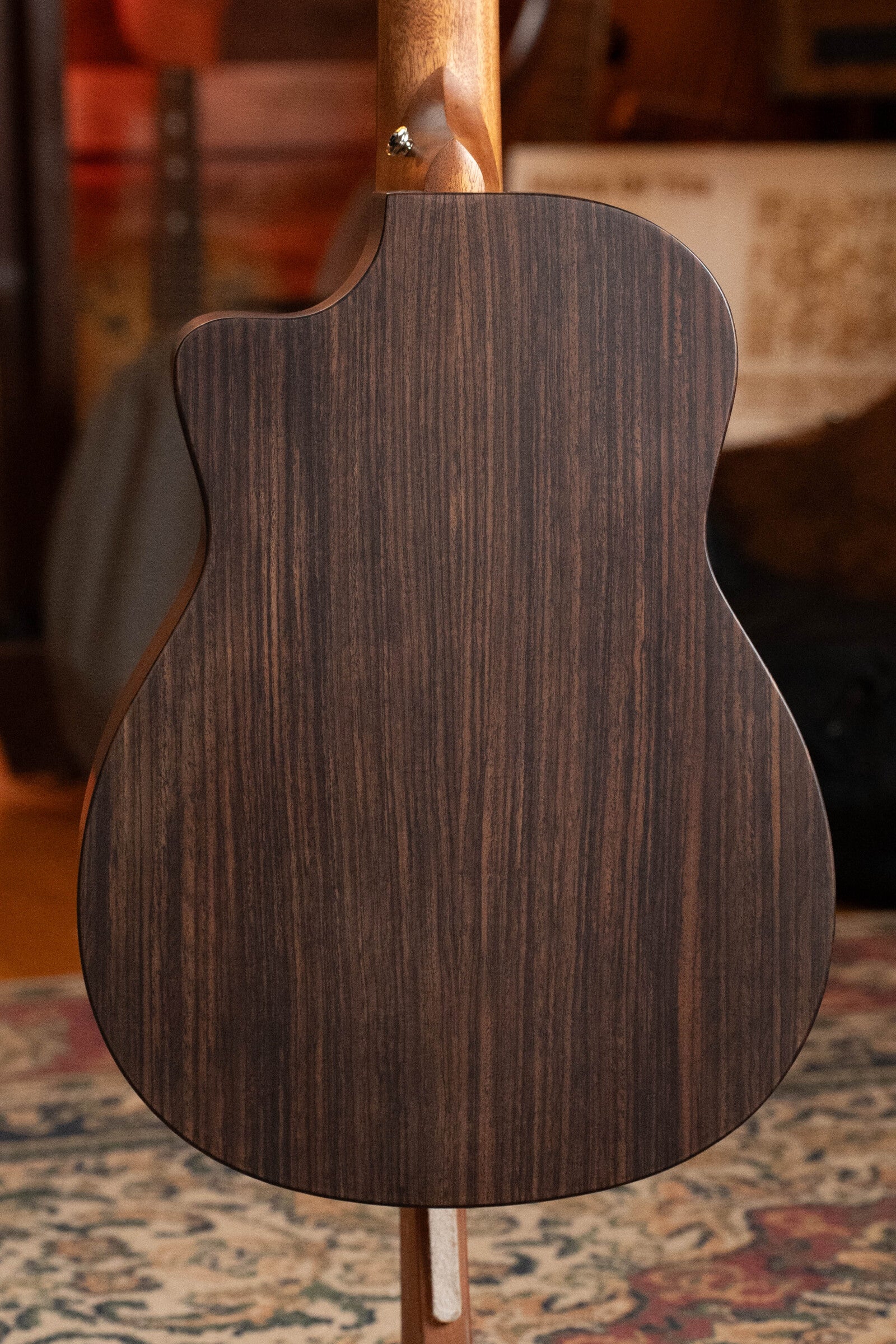 The Furch Pioneer-ER VTC travel guitar, crafted with Engelmann spruce and Indian rosewood, stands on a patterned rug in a warmly lit room, its rich dark wood grain back visible and musical gear in the background.