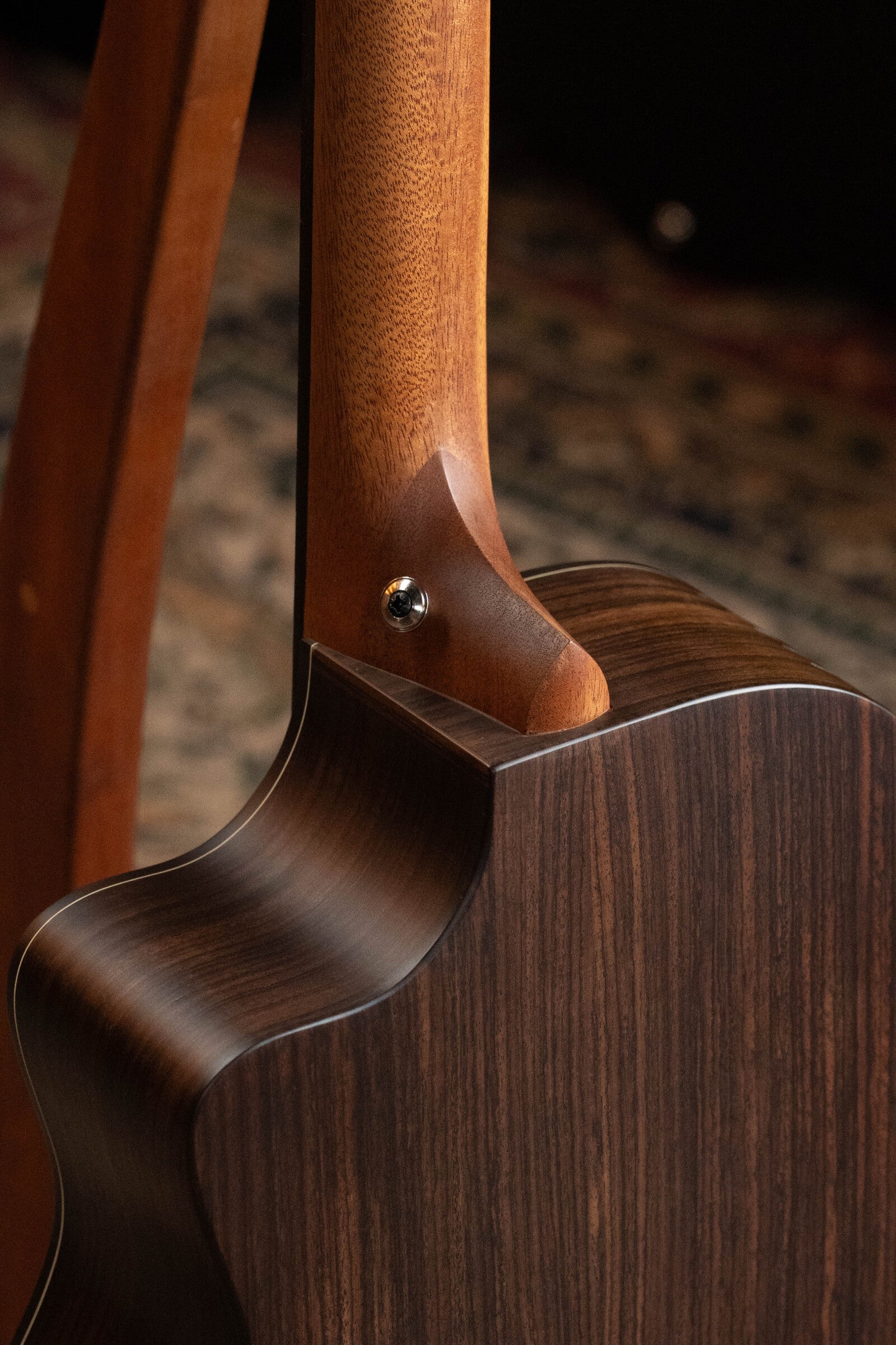 Close-up of the Furch Pioneer-ER VTC Engelmann Spruce/Indian Rosewood Cutaway Travel Guitar's neck joint and back, highlighting smooth wood grain and craftsmanship. The guitar, featuring Soundport Boosters & VTC Pickup, rests on a stand over a patterned rug.