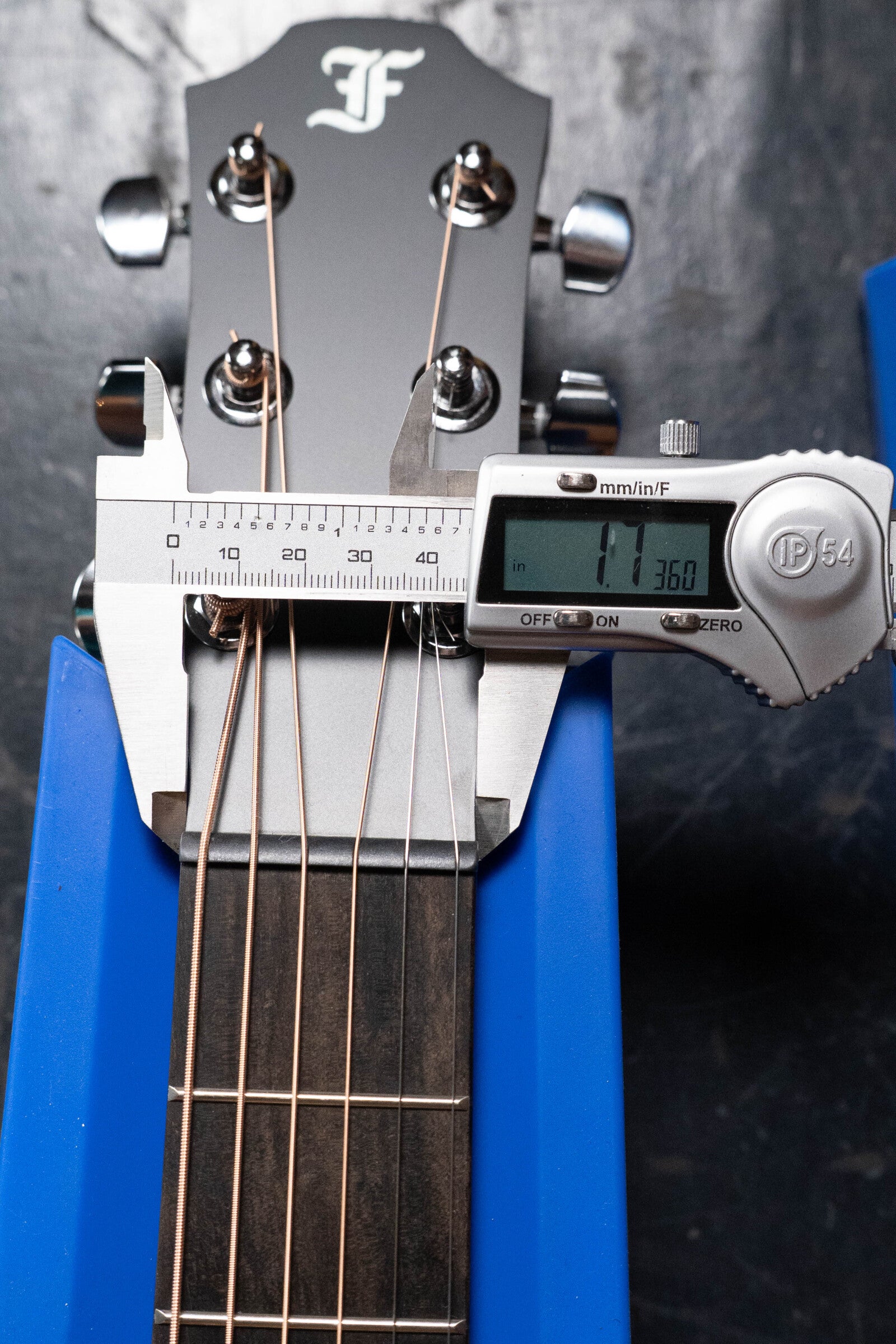 A digital caliper measures the nut width of a Furch Pioneer-ER VTC Engelmann Spruce/Indian Rosewood Cutaway Travel Guitar, displaying 1.360 inches. The guitar rests on a blue stand with its fretboard clearly visible.